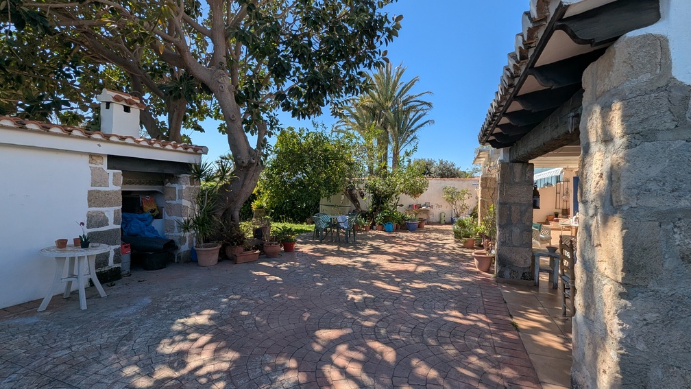 Spacious paved patio featuring traditional Tosca stone pillars, a shaded naya, and a summer kitchen outbuilding surrounded by mature trees and Mediterranean plants.