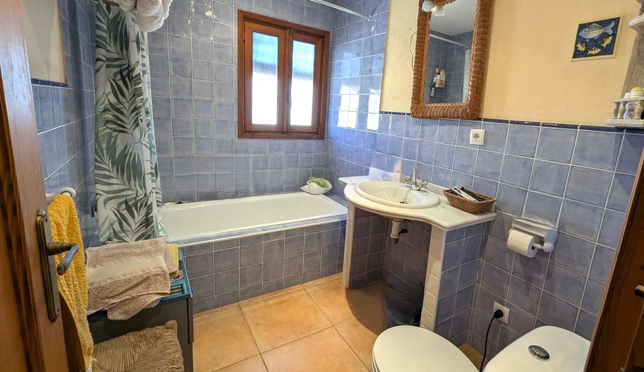 Traditional bathroom featuring blue ceramic wall tiles, a built-in bathtub, and a marble-topped vanity. A wooden-framed window provides natural light over the terracotta flooring.