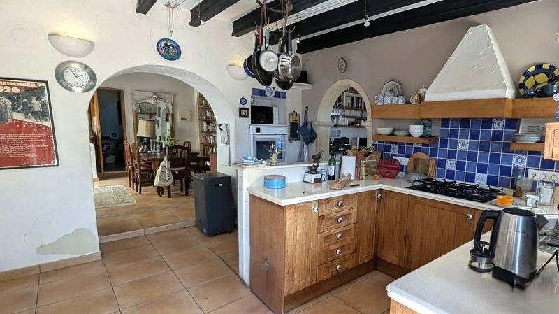 Traditional kitchen featuring wooden cabinetry, blue tiled backsplash, and dark ceiling beams. Arched openings lead to the adjacent dining area with terracotta flooring.