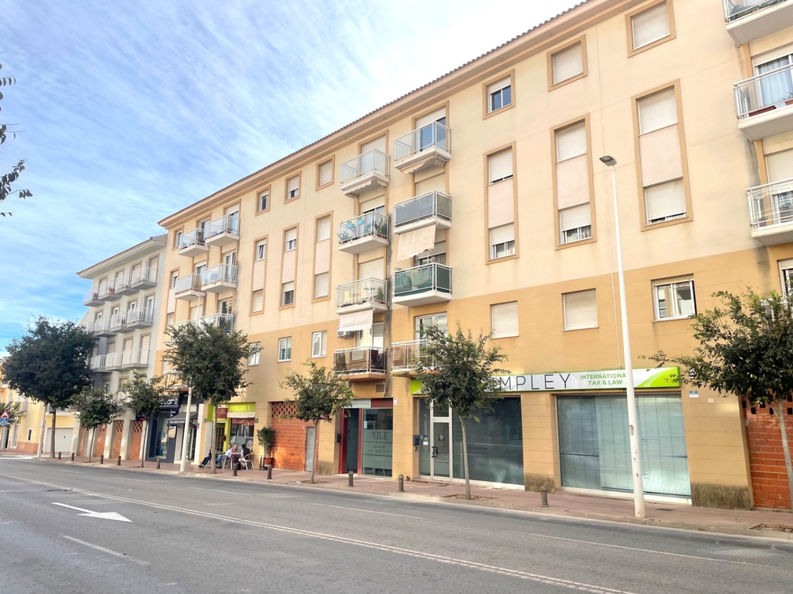 Exterior facade of a multi-story residential building featuring ground-floor commercial units, glass balconies, and a tree-lined sidewalk on a main avenue.