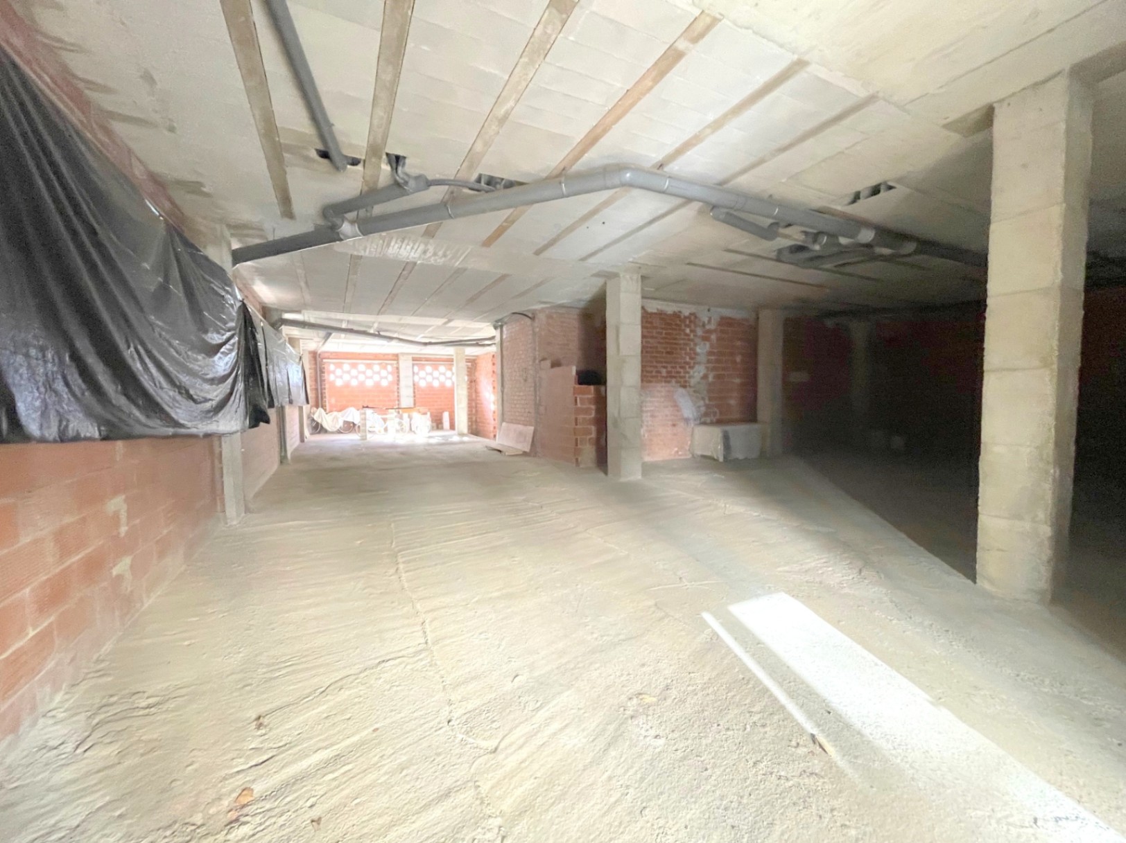 Open-plan commercial space in raw construction state, featuring concrete structural pillars, exposed ceiling pipes, and natural light from the rear.