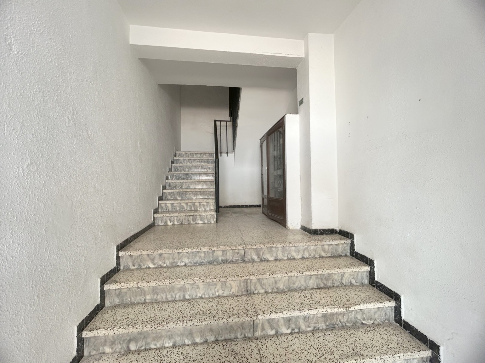 Common staircase featuring terrazzo steps with marbled edges, white walls, and black metal railings. A wooden display cabinet is positioned on the intermediate landing.