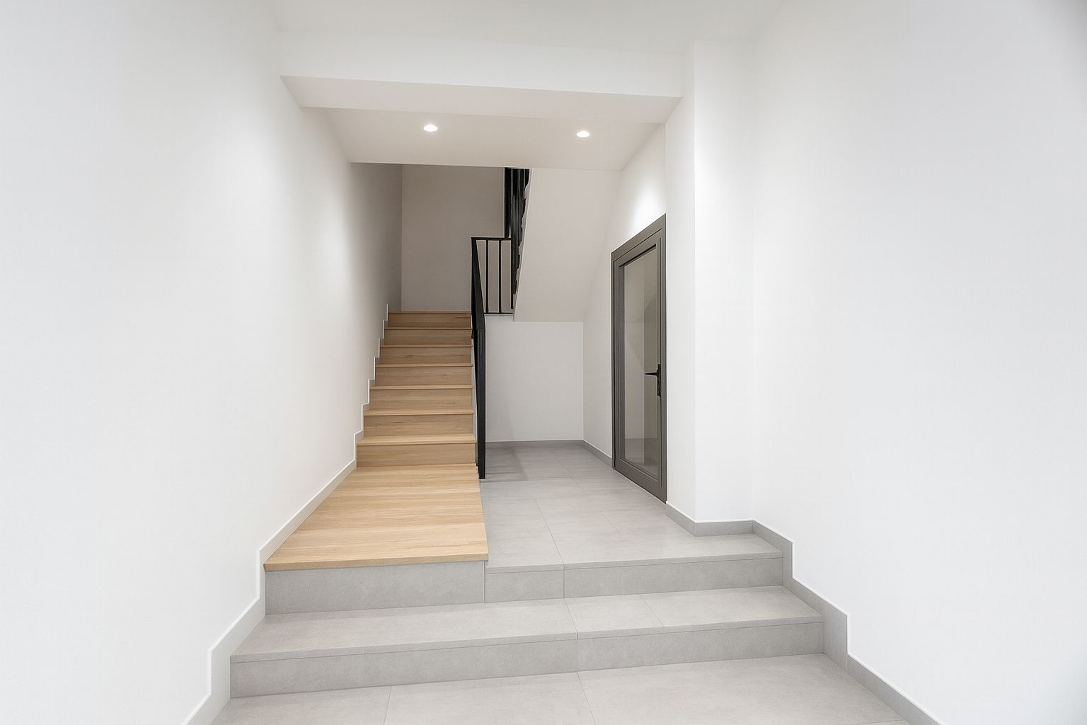 Entrance hallway featuring a light wood staircase with black metal railings. Grey tiled flooring, white walls, and a glass door with a grey frame.