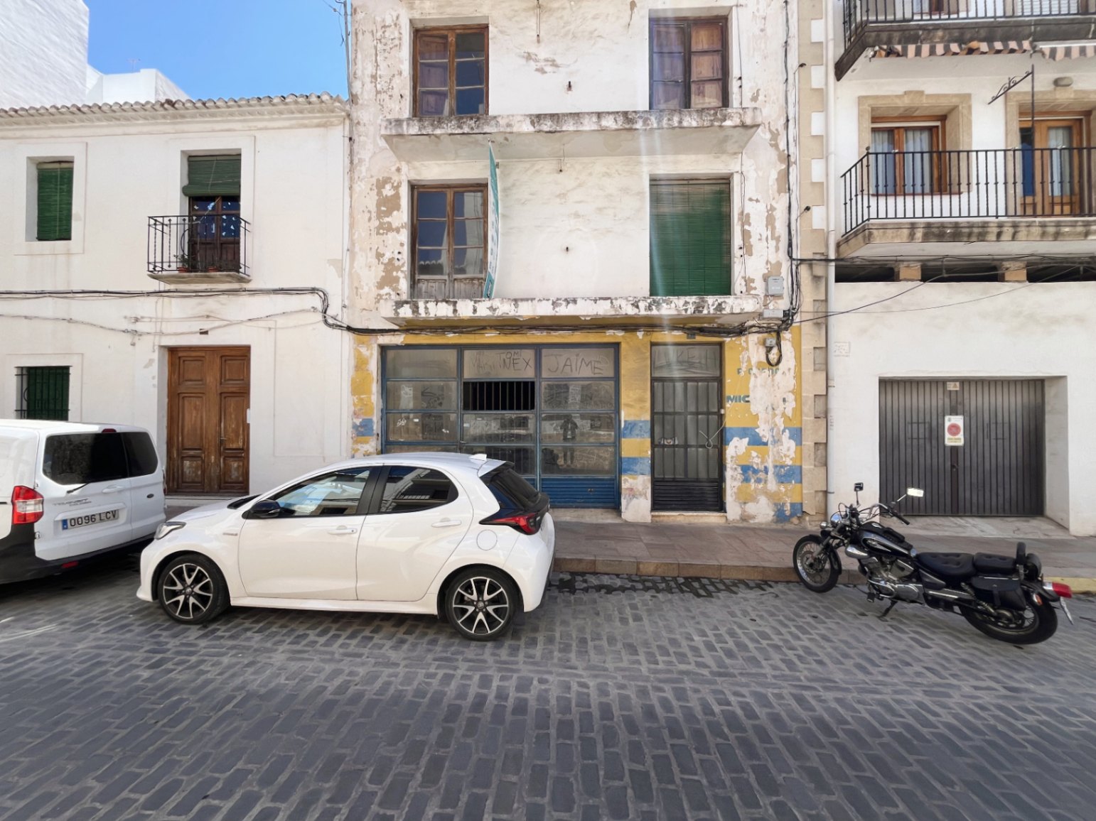 Exterior facade of the building in Jávea featuring a ground-floor commercial space, upper balconies, and wooden window frames on a traditional cobblestone street.