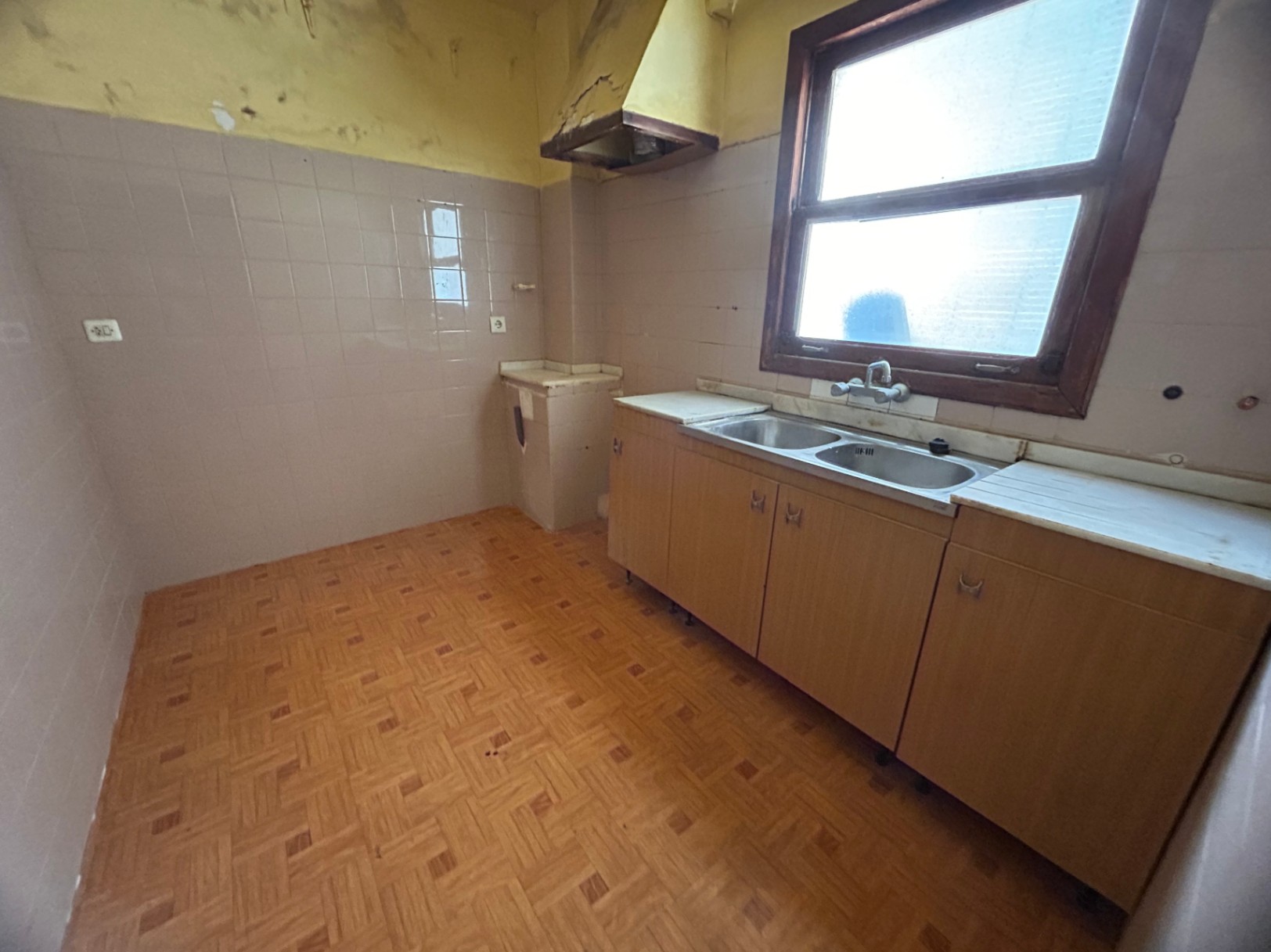 Original kitchen featuring a double stainless steel sink, wooden cabinetry, and patterned floor tiles. A wooden-framed window provides natural light for this renovation project.