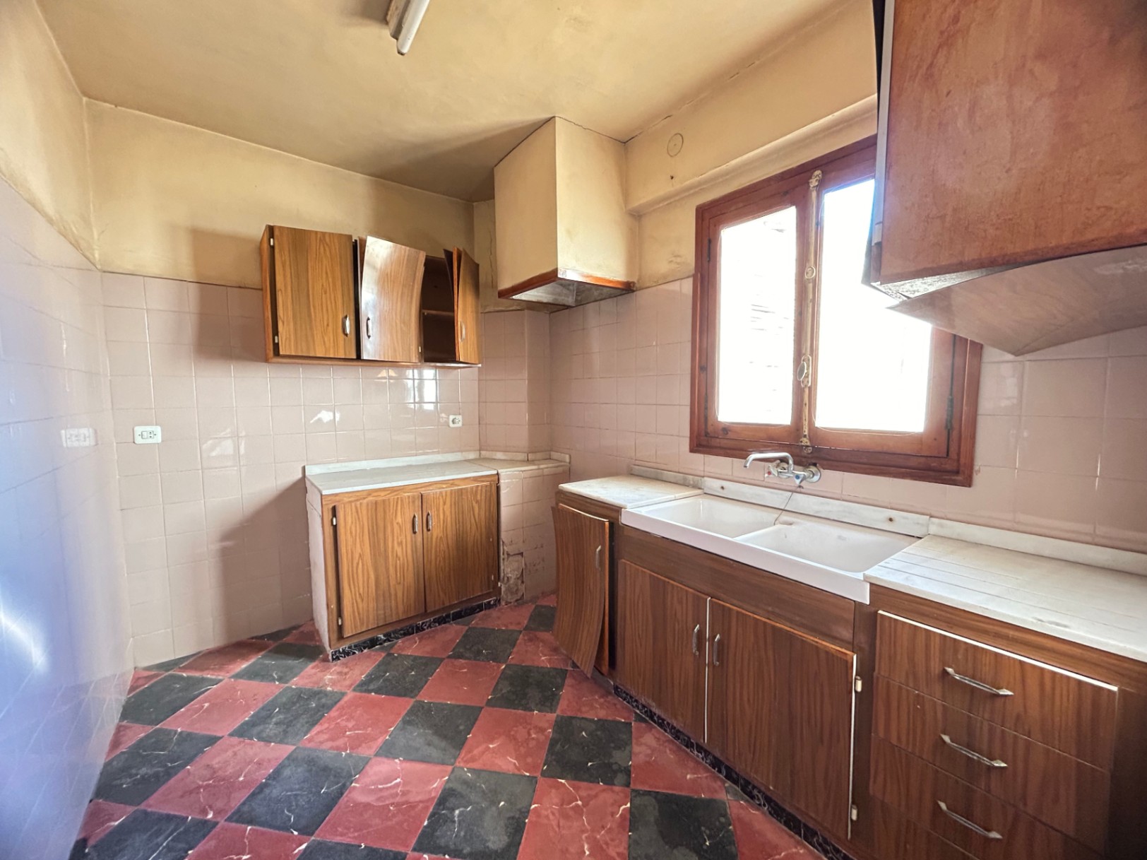 Original kitchen featuring red and black checkered tile flooring, wooden cabinetry, and white marble countertops. Includes a double basin sink and a wooden window frame.