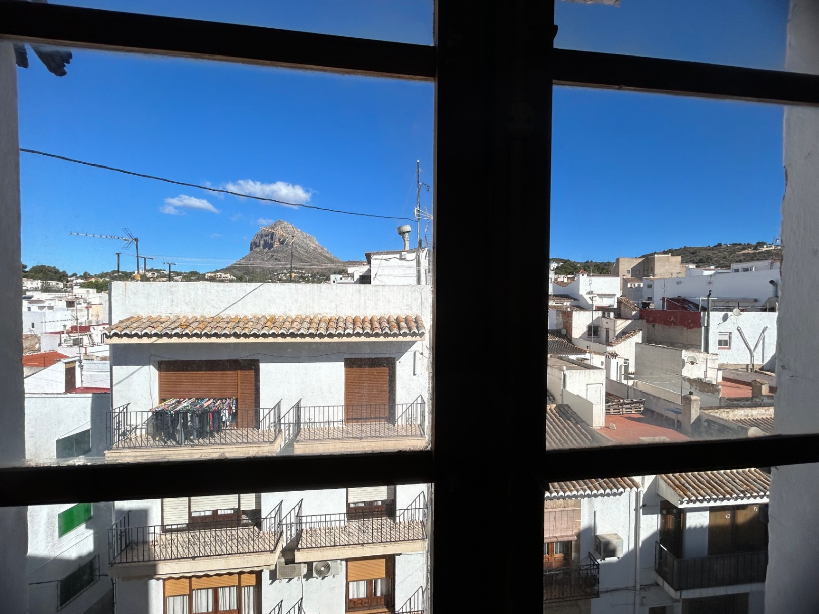 Urban view from indoors through a window frame, overlooking the rooftops of Jávea's old town with the Montgó mountain peak visible in the distance.