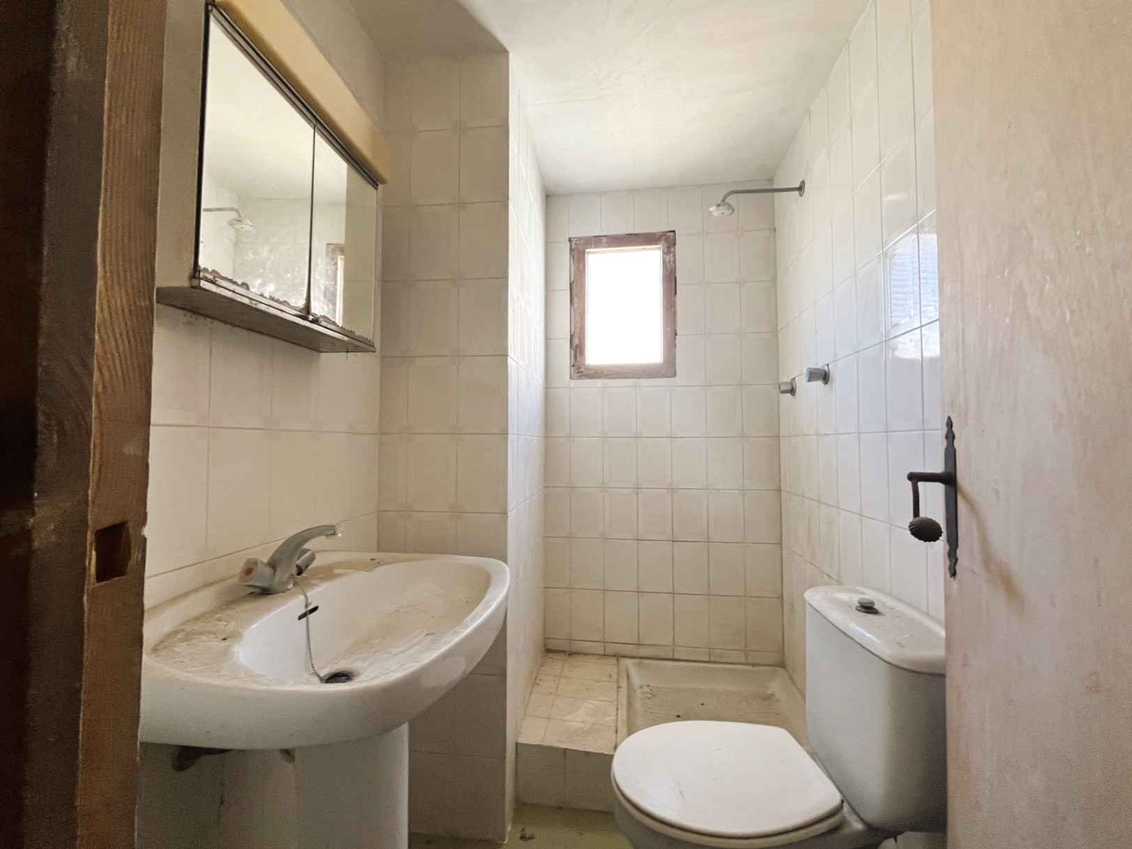 Functional bathroom featuring white tiling, a pedestal sink, and a shower area with a small window. A space with significant renovation potential.