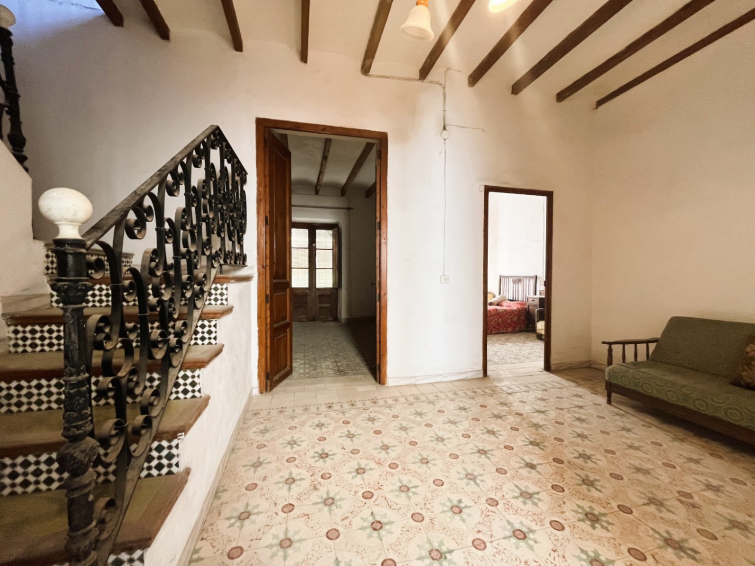Hallway featuring original hydraulic tile flooring, exposed wooden ceiling beams, and a grand staircase with an ornate wrought iron railing.