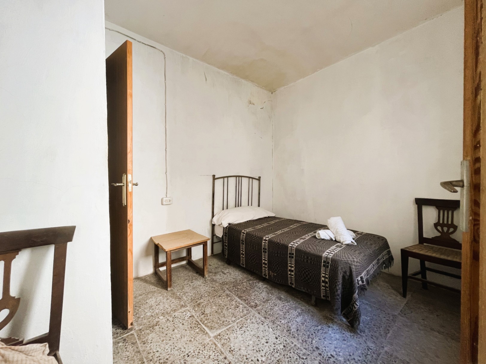 Simple bedroom featuring original terrazzo flooring, a single bed with a metal headboard, and traditional wooden furniture against white walls.