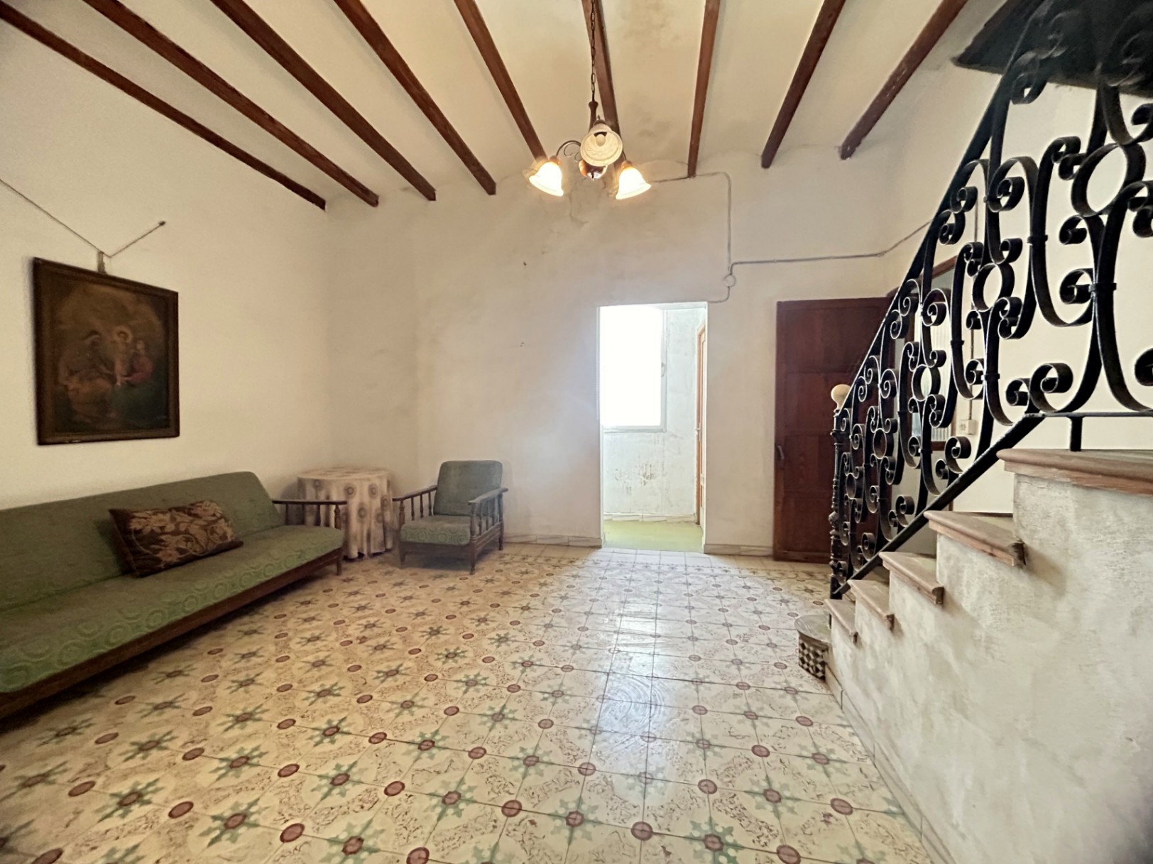 Stately living area featuring original hydraulic tile flooring, high ceilings with exposed wooden beams, and a staircase with ornate wrought iron railings.