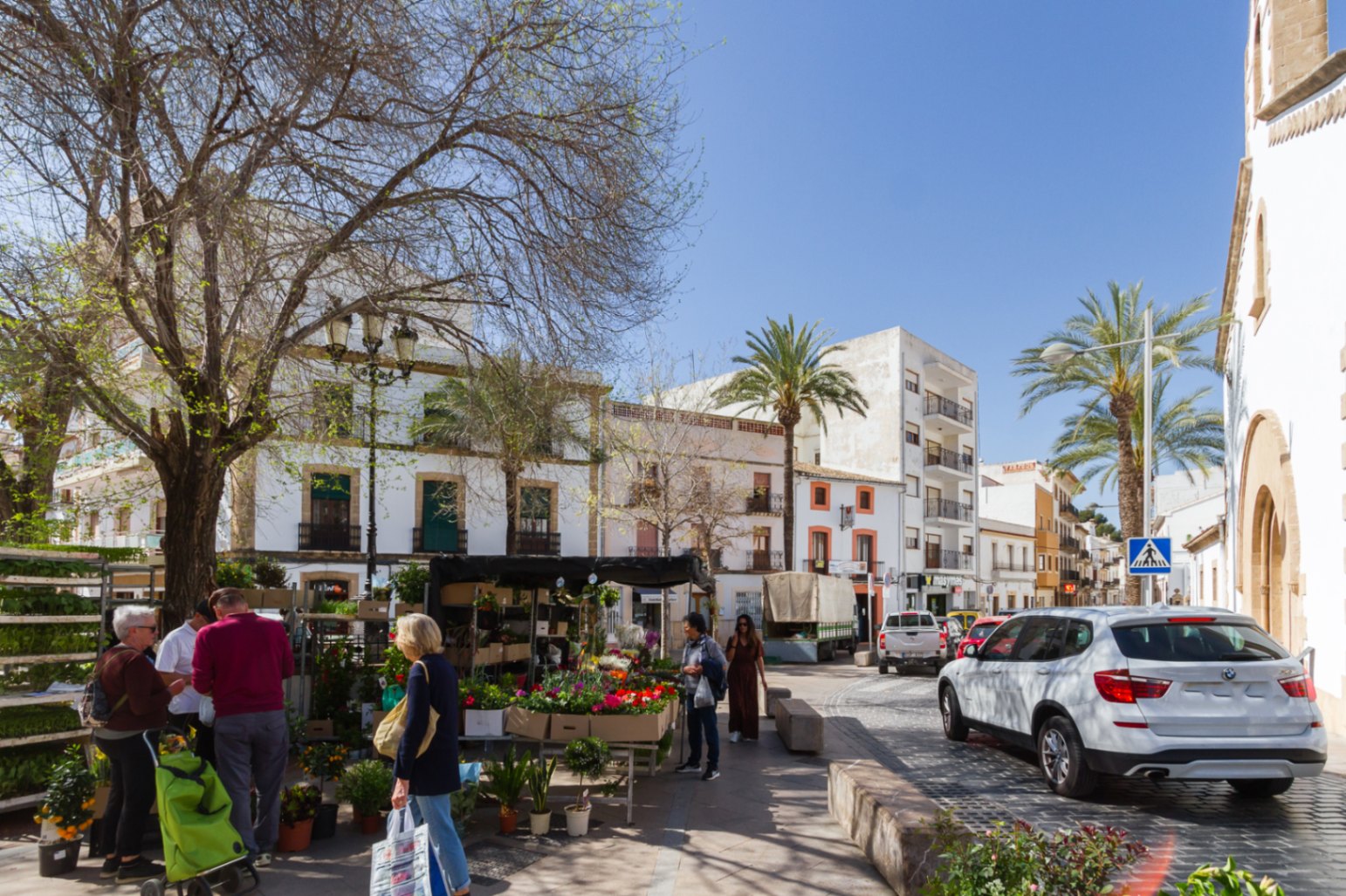 A square in Jávea's historic center featuring a local flower stall, traditional white-facade architecture, cobblestone streets, and palm trees under a clear sky.
