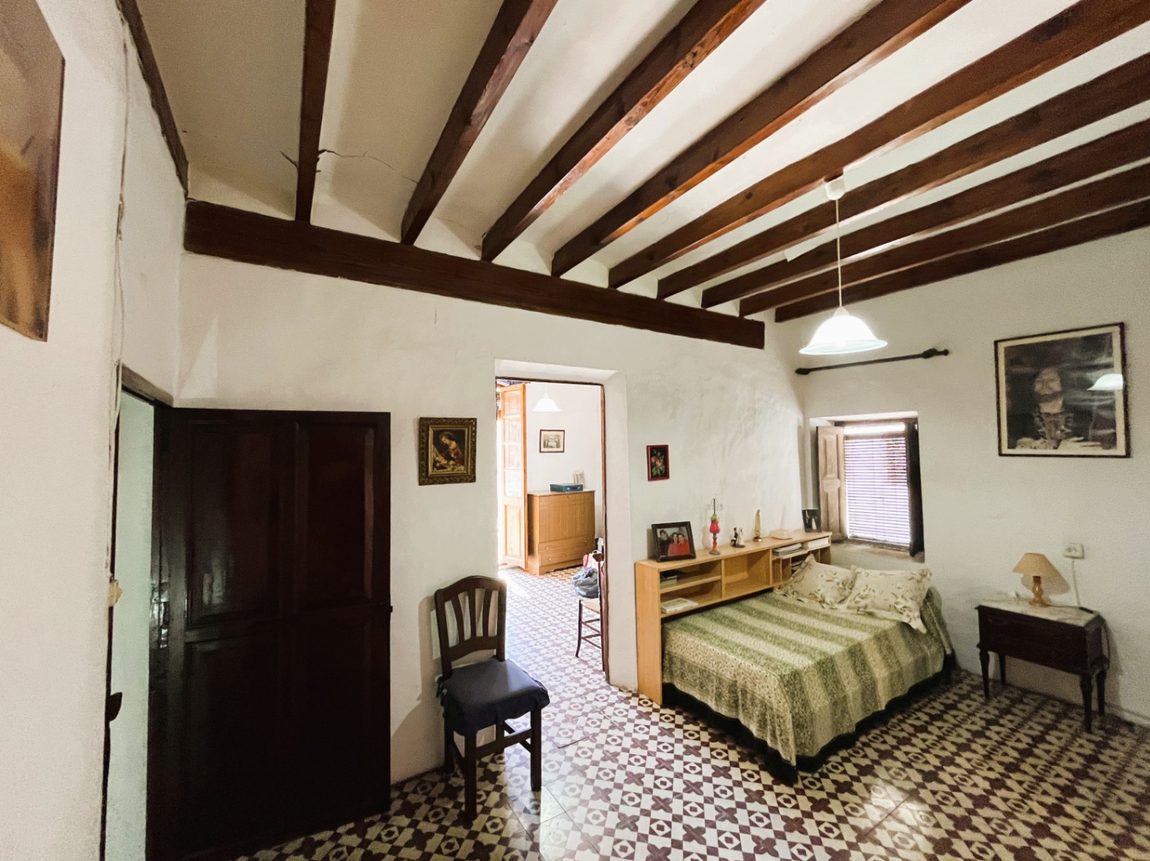 Bedroom featuring exposed dark wood ceiling beams, original hydraulic floor tiles, and thick white walls. It includes a small window with shutters and built-in wooden shelving.