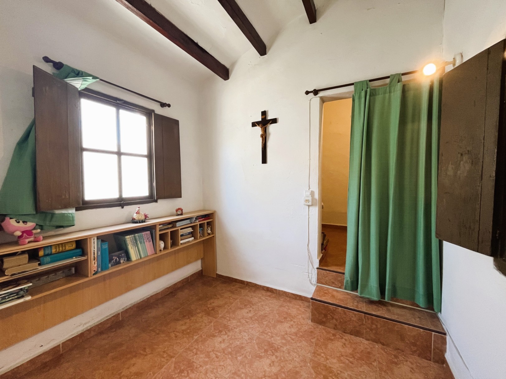 Interior space featuring exposed dark wood beams, terracotta flooring, and a low bookshelf. An elevated doorway with steps and a green curtain leads to another room.