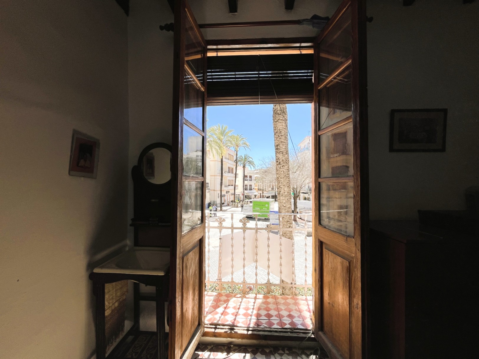 Interior view through traditional wooden doors opening onto a balcony with red checkered tiles, overlooking a sunlit plaza with palm trees in the old town.
