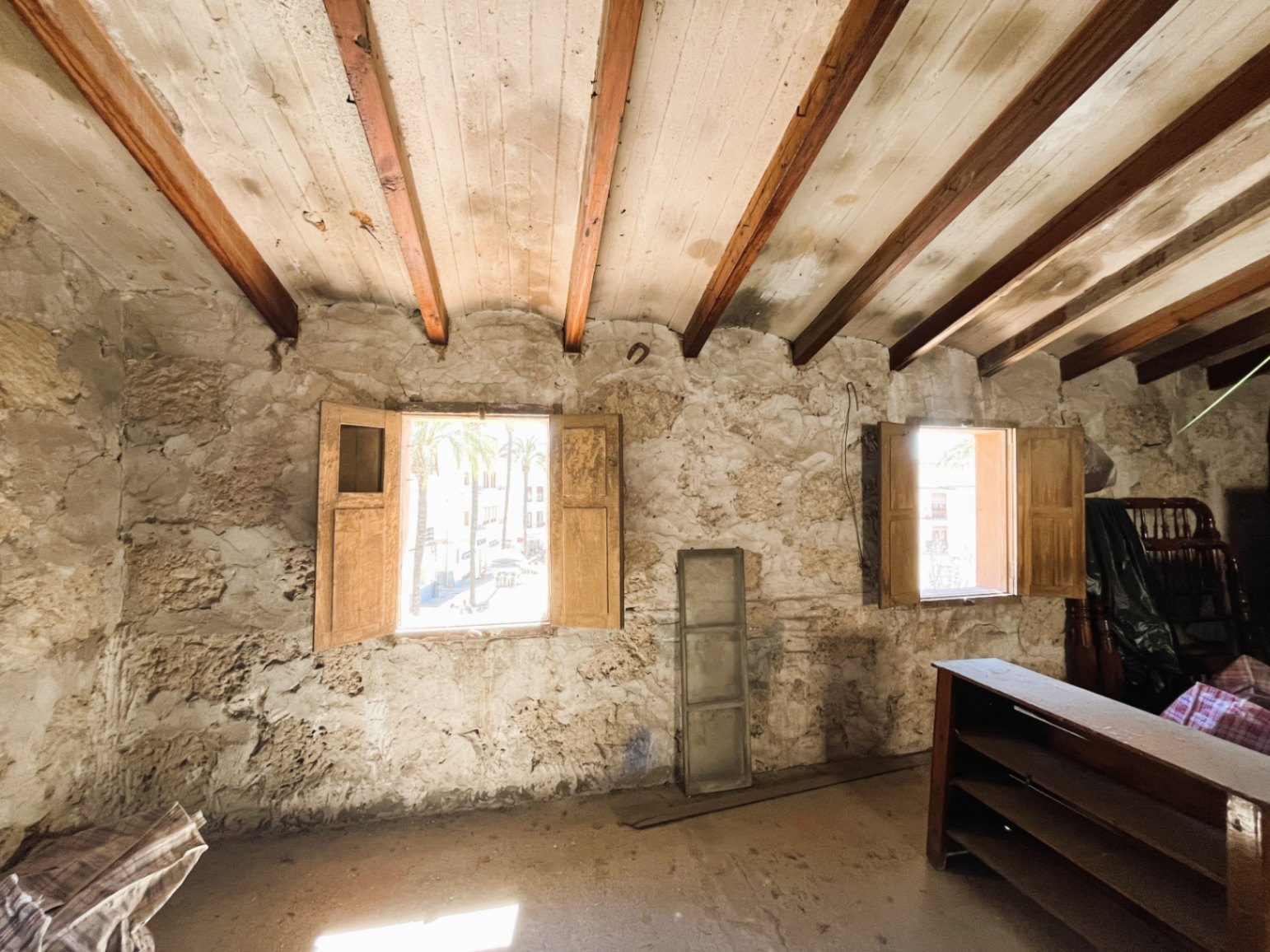 Room featuring original exposed stone walls and traditional wooden ceiling beams. Two windows with wooden shutters provide natural light and views of the historic town.