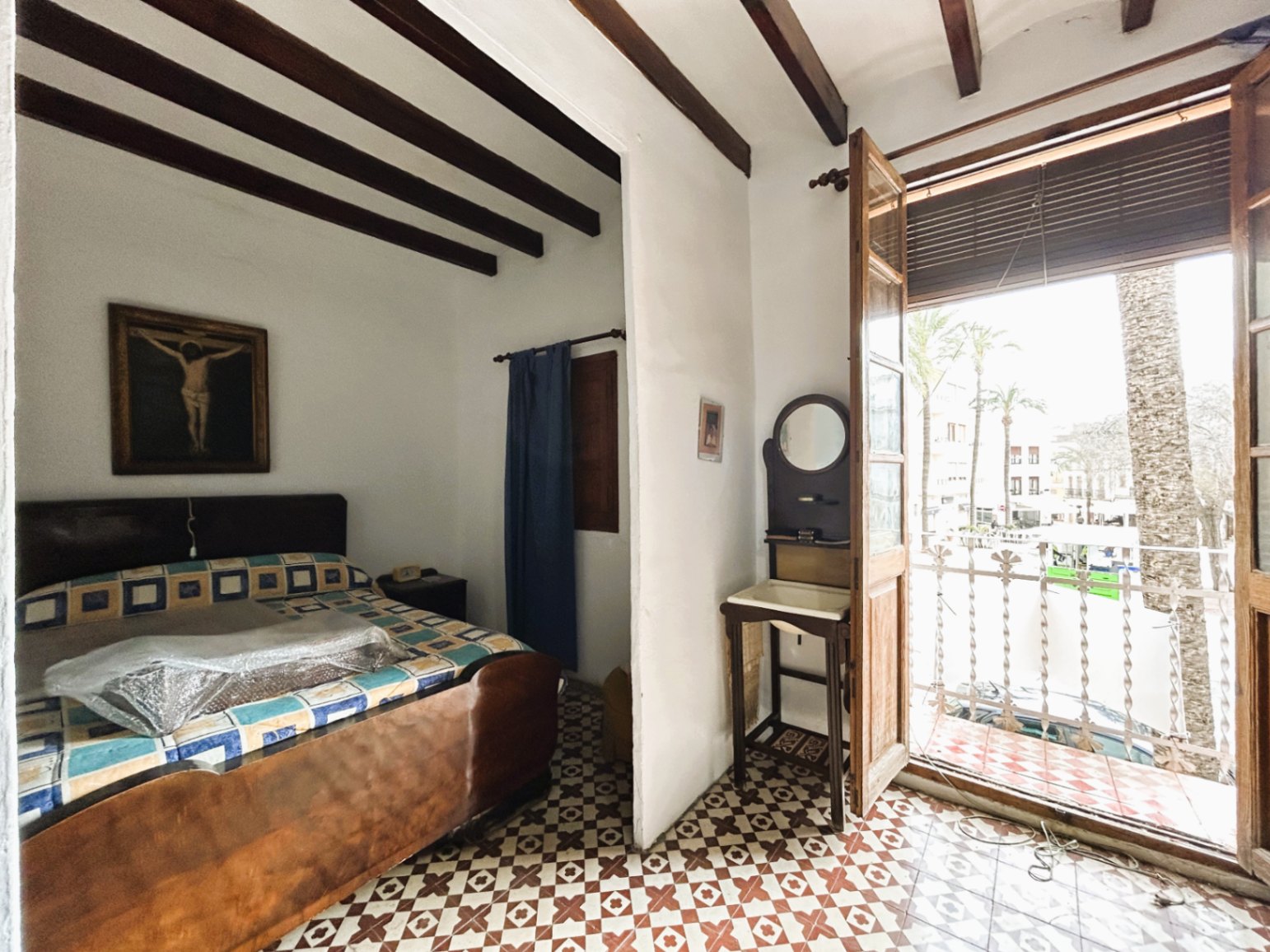 Traditional bedroom featuring exposed wooden ceiling beams, geometric hydraulic floor tiles, and a balcony overlooking the town square and palm trees.
