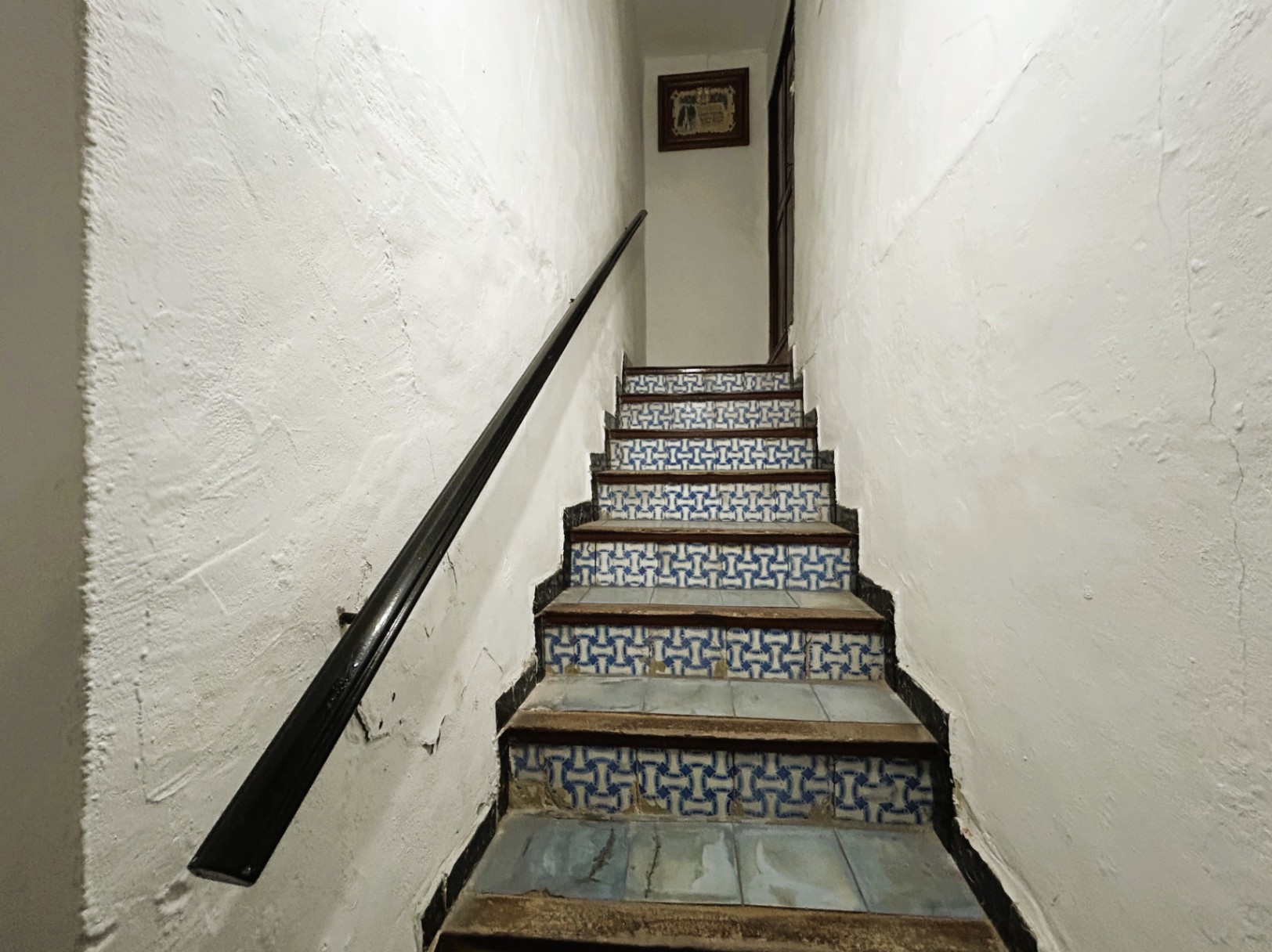 Traditional staircase featuring blue and white patterned tile risers, wooden treads, and a dark handrail set against textured white plaster walls.