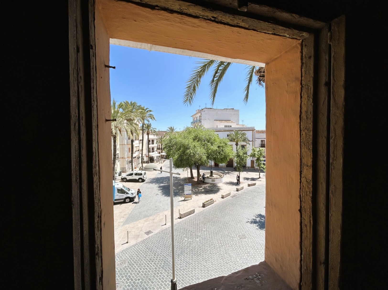 View from the interior overlooking a cobblestone square in the old town, showcasing traditional thick masonry walls and a sunlit plaza with palm trees.