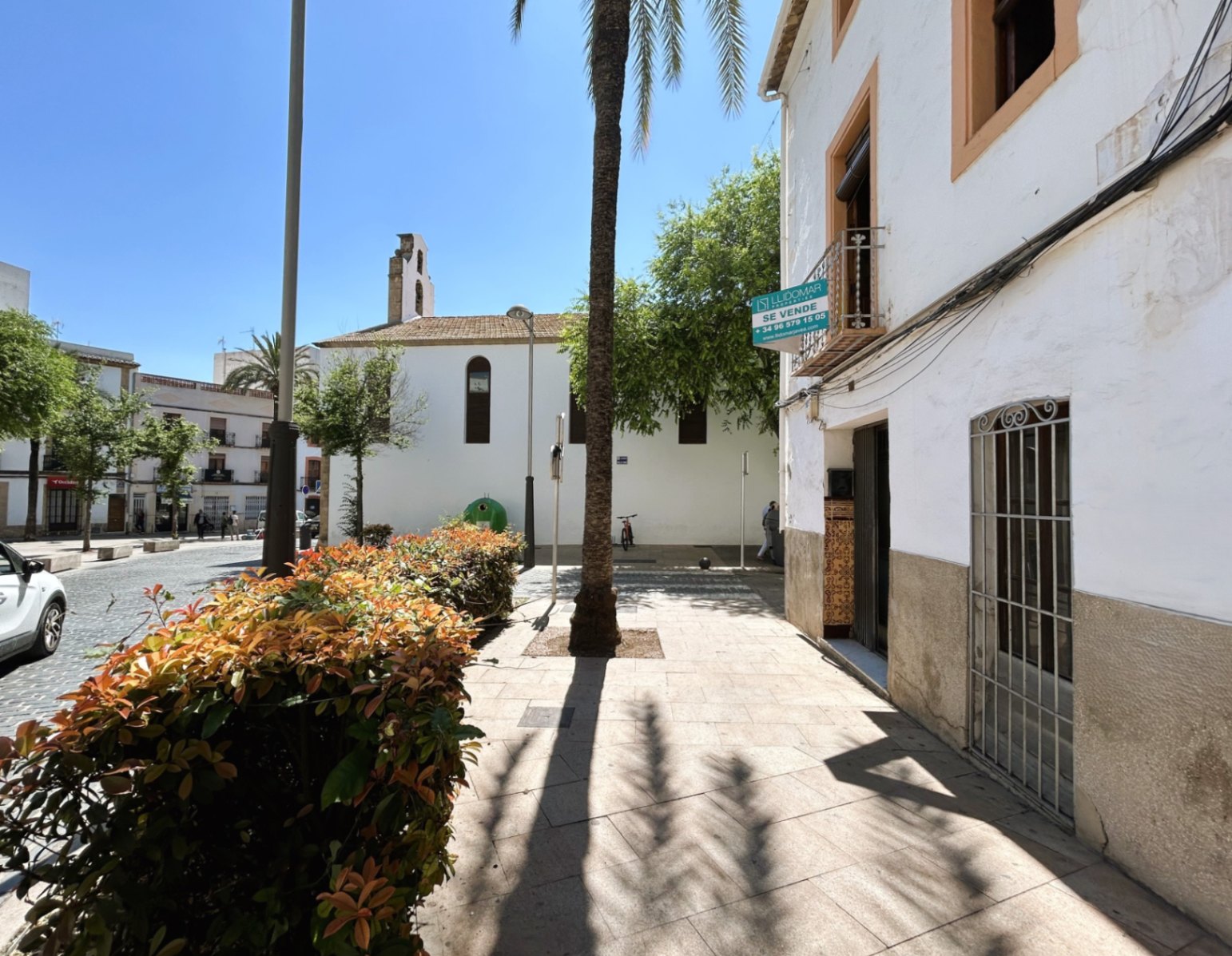White facade with a stone base in Jávea's old town, located next to a pedestrian plaza with palm trees and views of a historic bell tower.