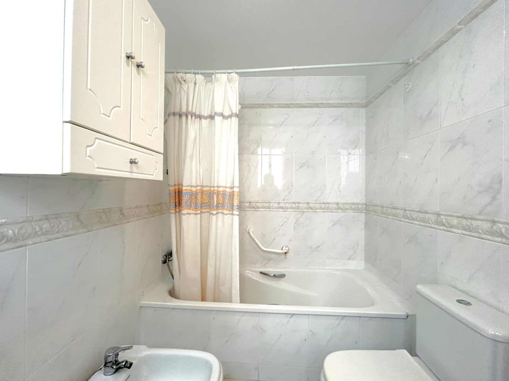 Bathroom featuring a built-in bathtub with marble-effect tiling and decorative borders. Includes a bidet, toilet, and white wall-mounted storage cabinetry.