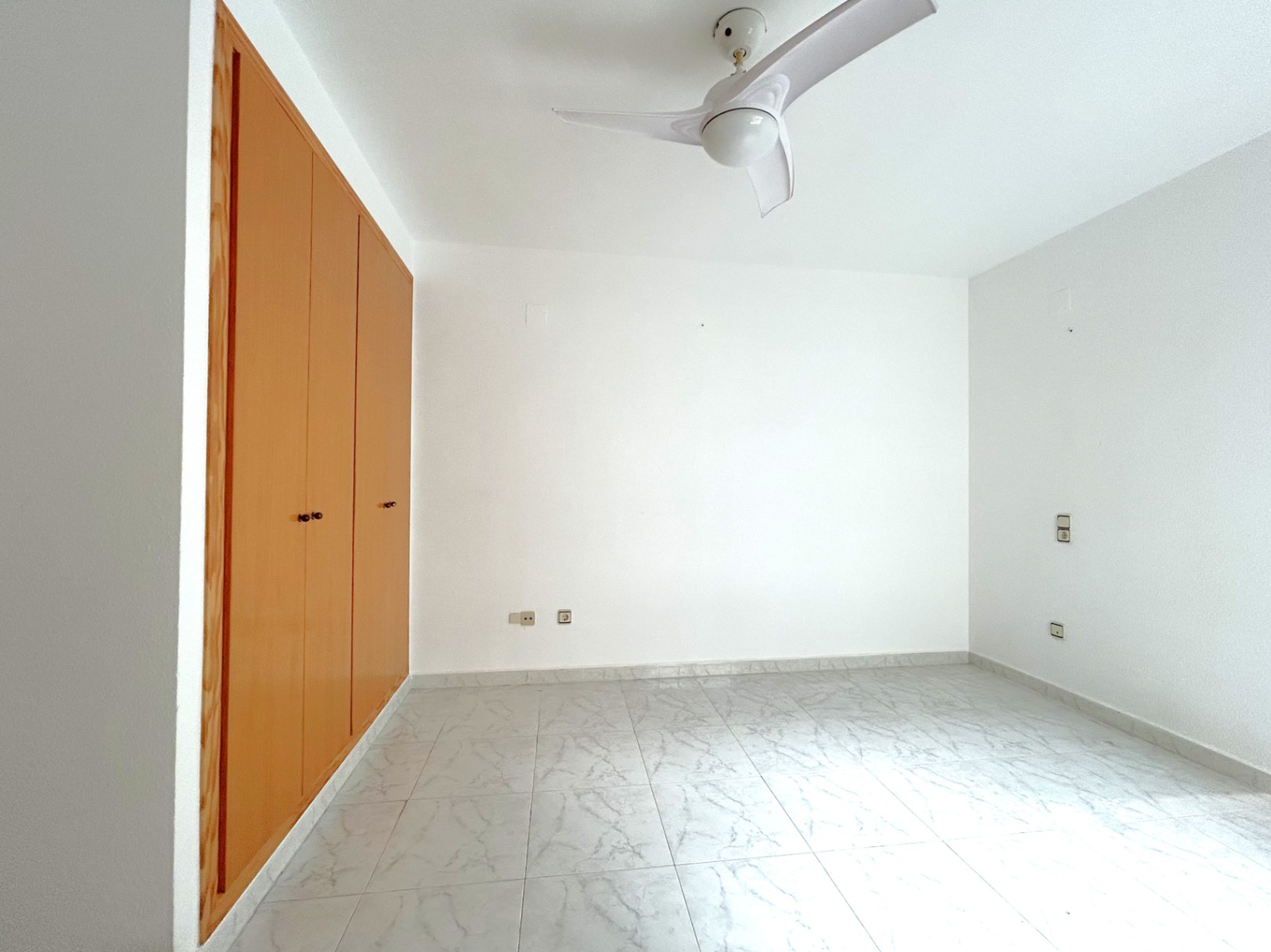 Bright bedroom featuring a large built-in wooden wardrobe, marble-effect floor tiles, and a modern white ceiling fan.