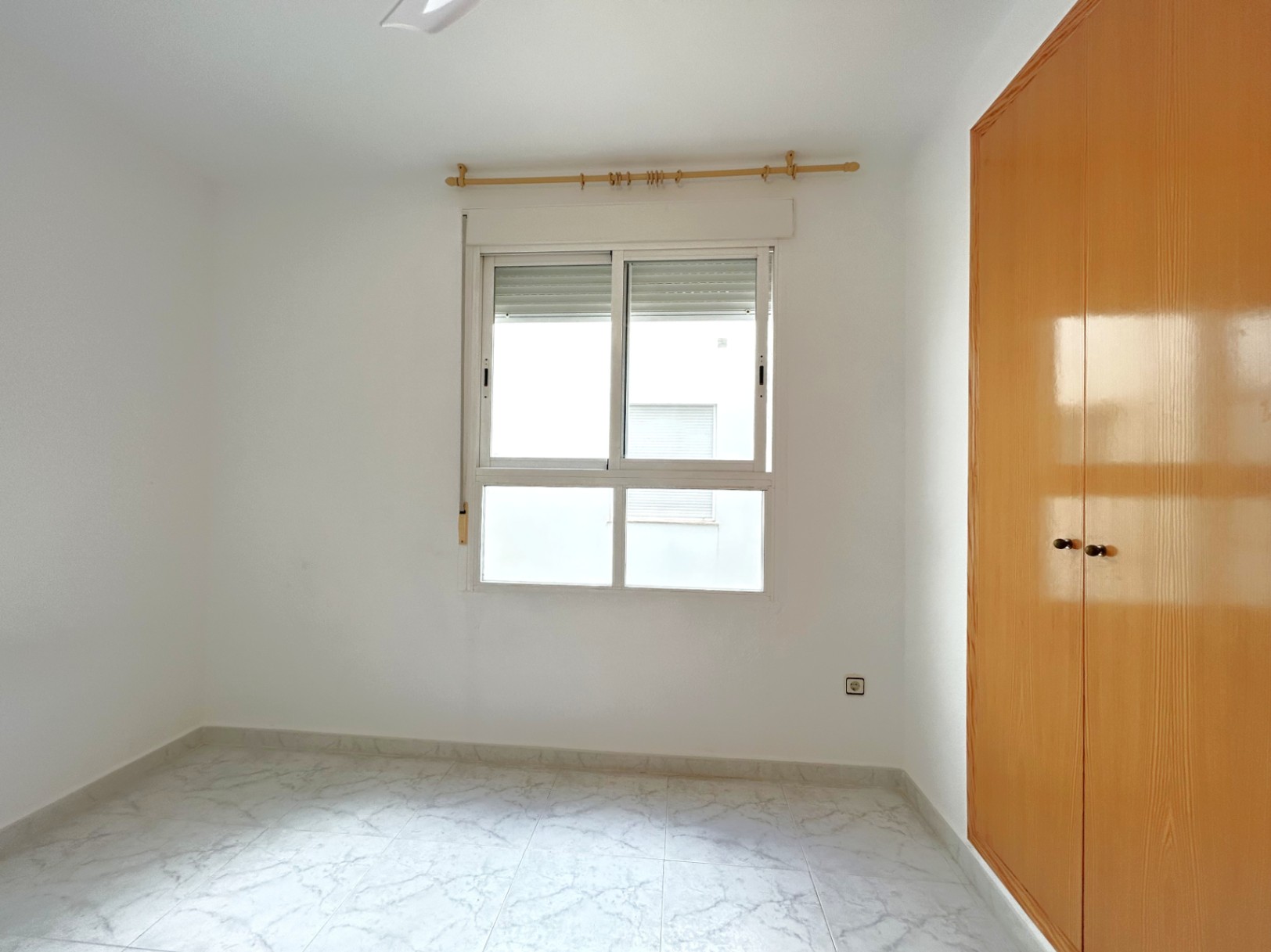 Bright bedroom featuring marble-effect floor tiles, a built-in wooden wardrobe, and a white aluminum window with integrated shutters.