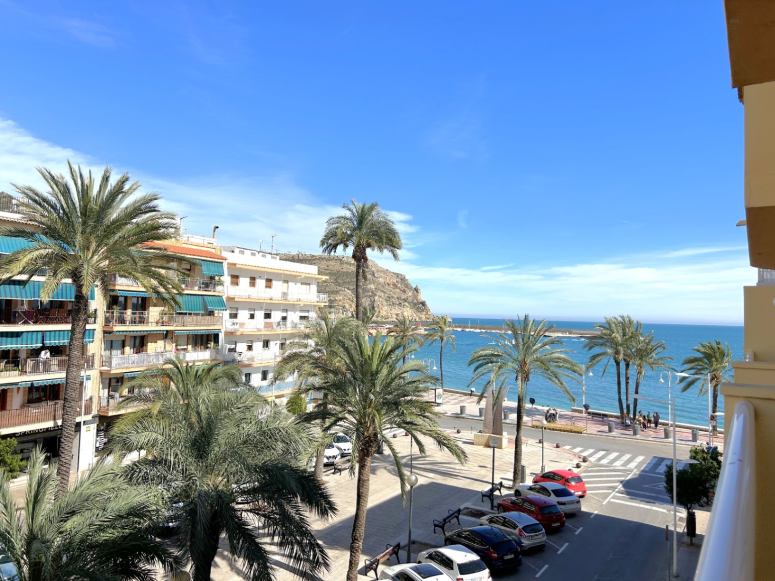 Elevated balcony view overlooking the Jávea Port promenade, featuring palm trees, the Mediterranean Sea, and coastal cliffs under a clear sky.
