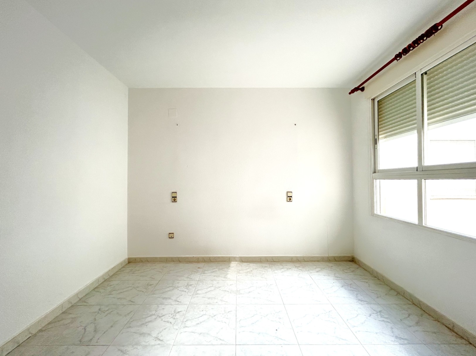 Empty bedroom featuring marble-effect ceramic flooring, white walls, and a window with integrated shutters. Symmetrical electrical outlets are positioned for a bed headboard.