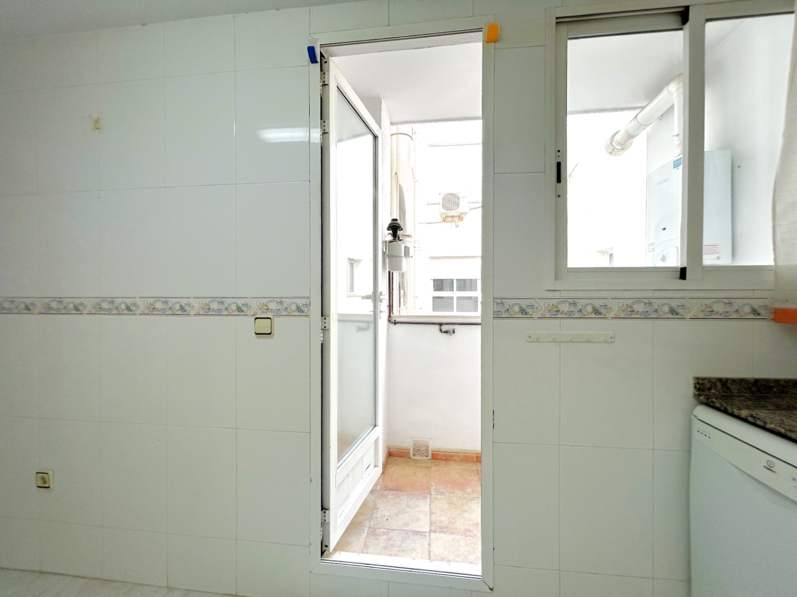 Exterior utility gallery accessed from the kitchen, featuring white tiled walls, granite countertops, and a laundry area with a water heater.