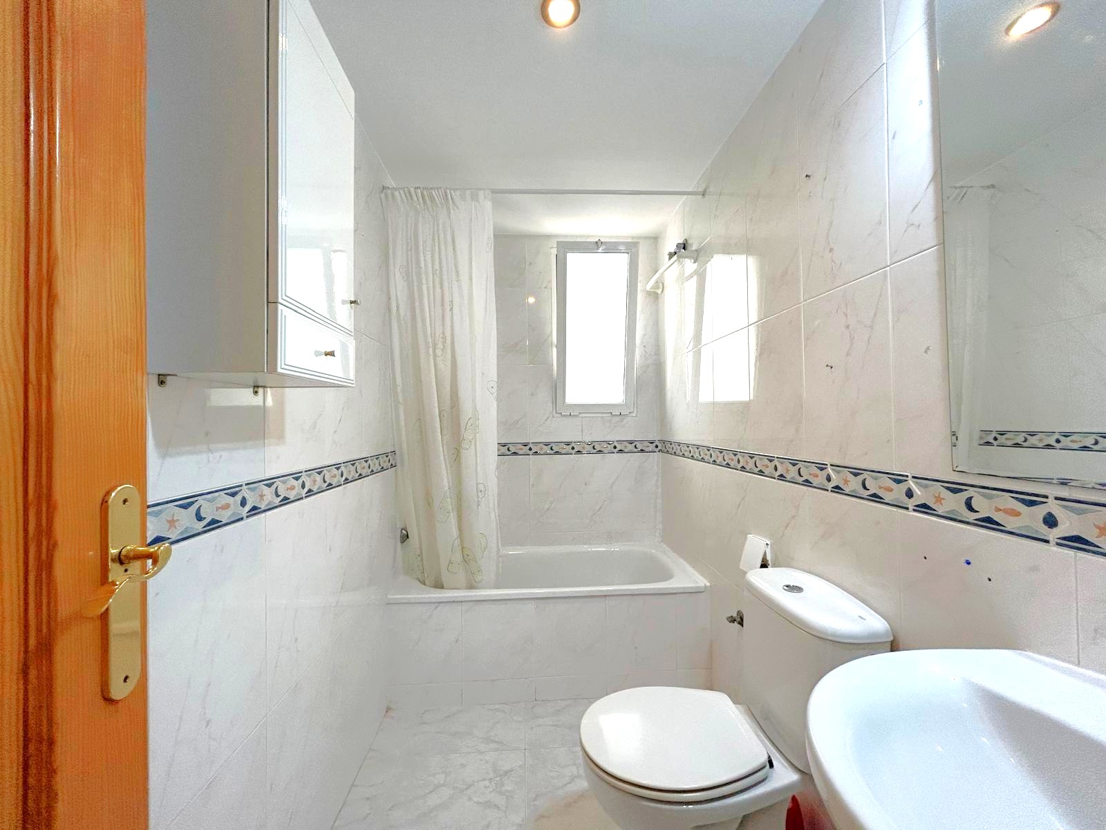 Full bathroom featuring a bathtub with a white curtain, marble-effect tiling with a decorative border, and a window providing natural light and ventilation.