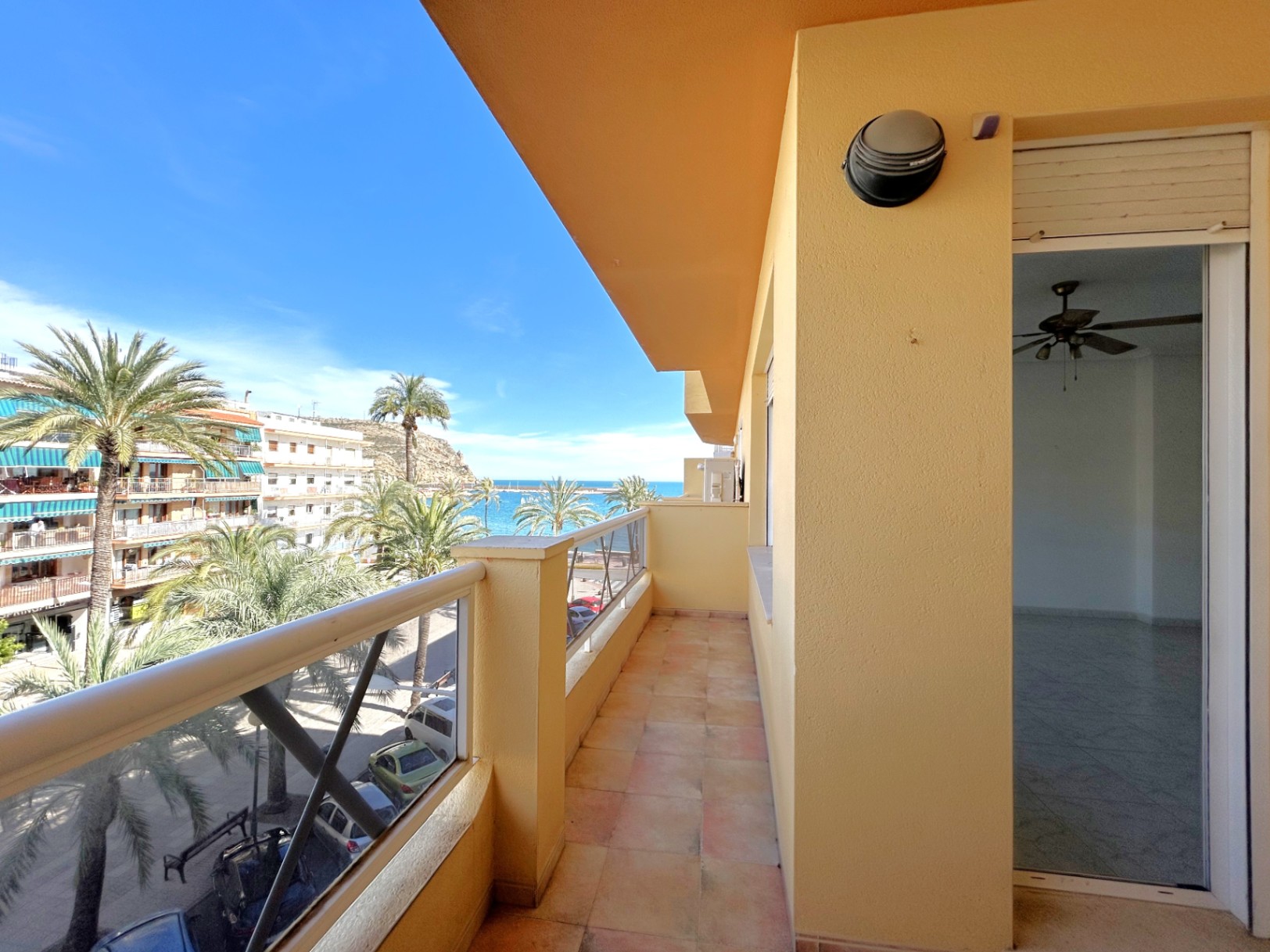 Long balcony with ceramic tiling featuring side views of the Mediterranean Sea and Jávea port, overlooking palm trees and the street.