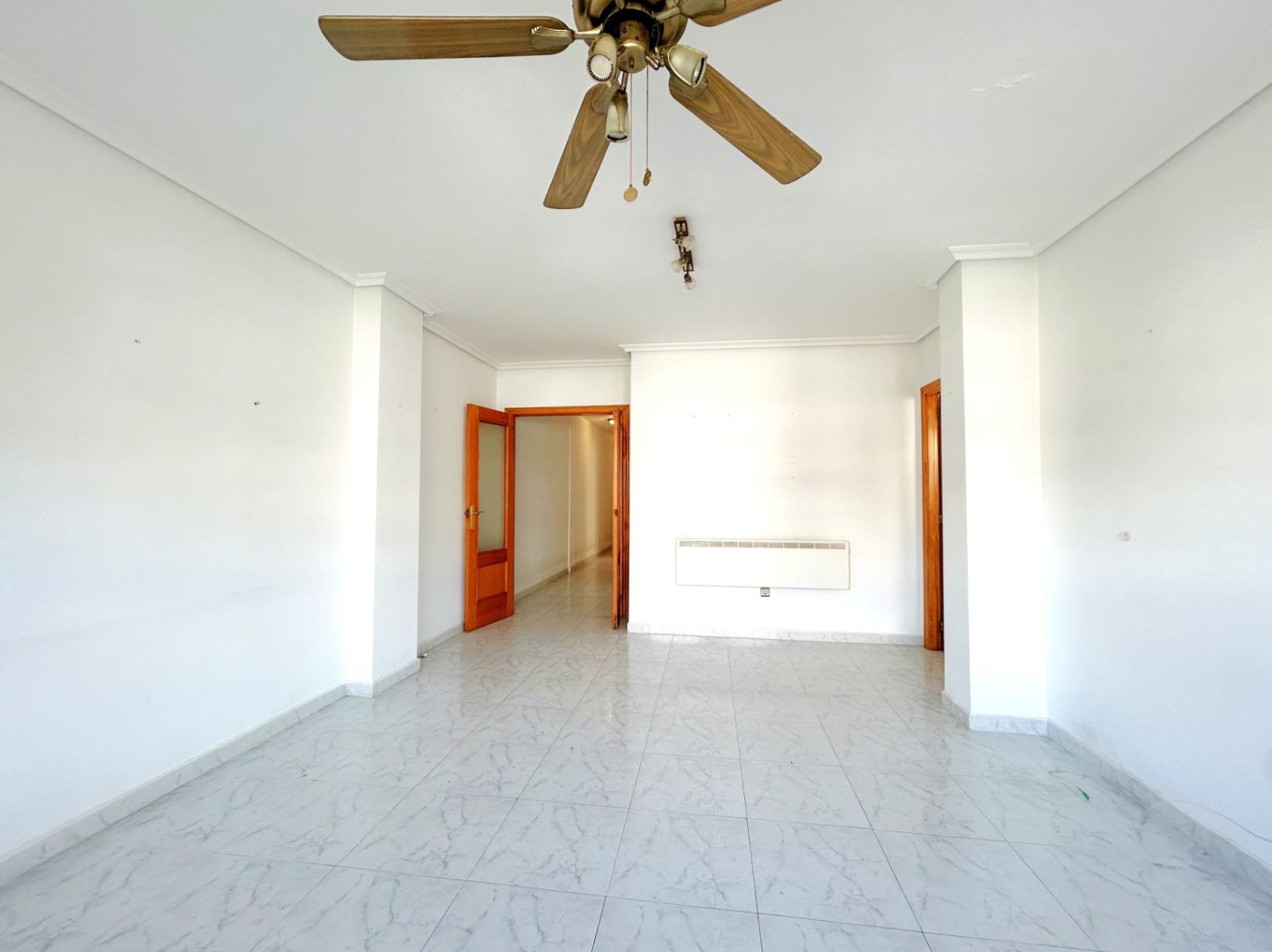 Spacious living area featuring marble-effect tiled flooring, white walls, and a wooden ceiling fan. The room includes wall-mounted heating and natural wood door frames.