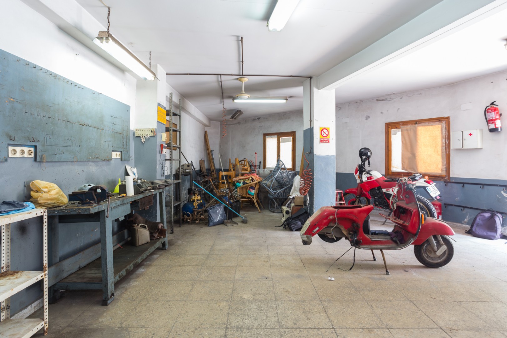 Spacious workshop or garage area featuring a workbench, metal shelving, and tiled flooring, equipped with fluorescent lighting and ceiling fans.