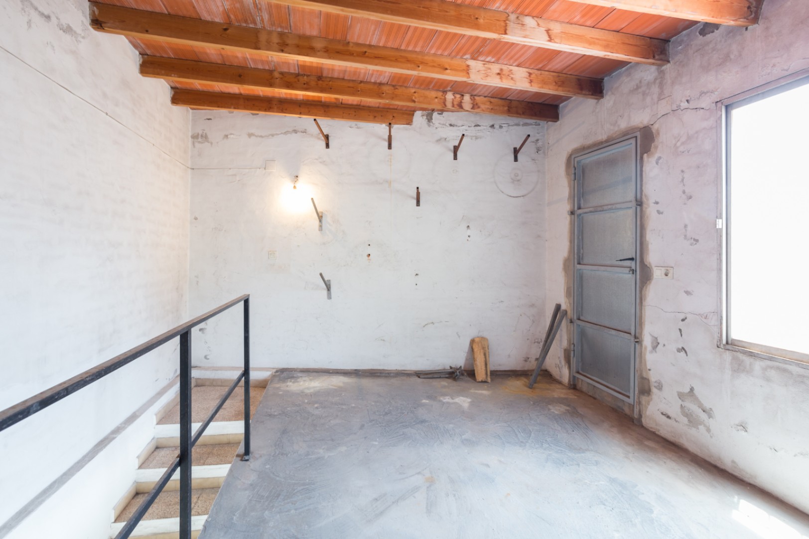 Upper floor featuring exposed wooden beams and terracotta ceiling tiles. The space includes a concrete floor, black metal staircase railing, and industrial-style metal door.