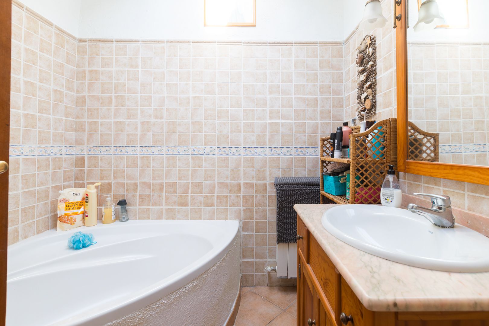 Bathroom featuring a corner bathtub, marble-topped wooden vanity, and walls finished with beige square tiles and a decorative blue border.