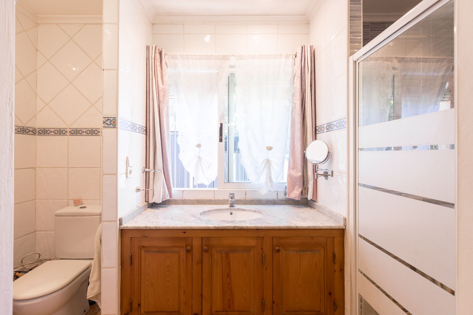 Bathroom featuring a wooden vanity with marble countertop, a central window with curtains, and a glass shower screen with frosted horizontal stripes.