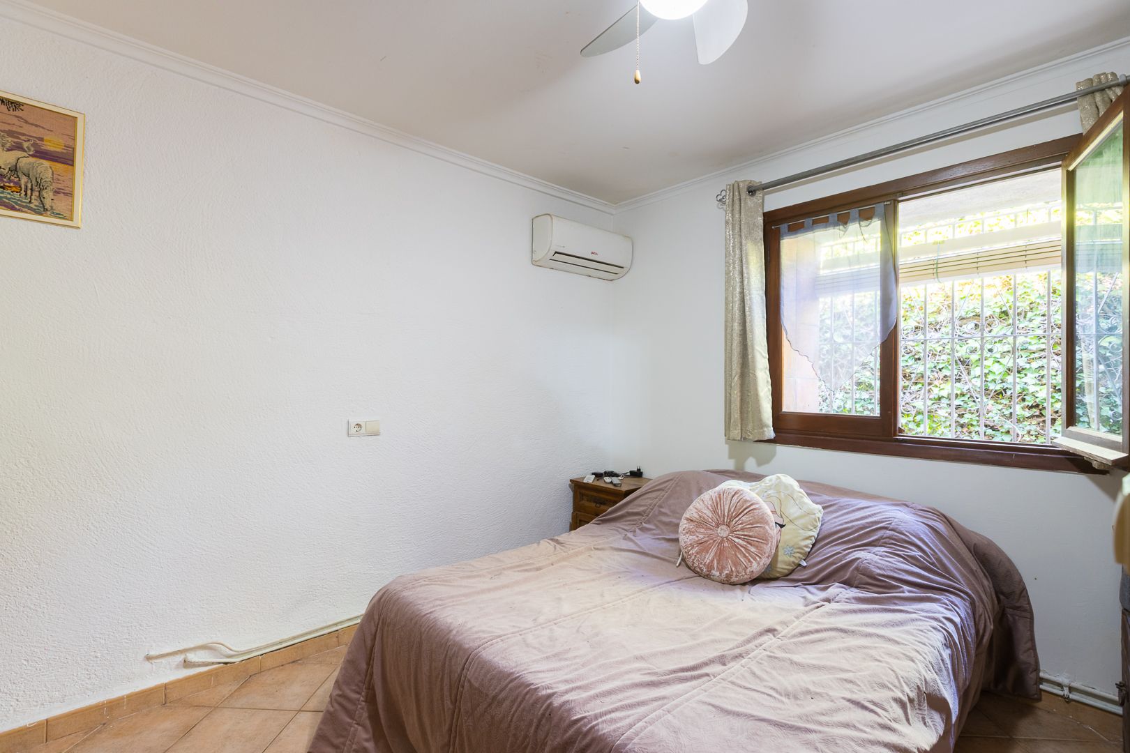 Bright bedroom featuring a split air conditioning unit, ceiling fan, and a wooden-framed window overlooking the garden greenery.