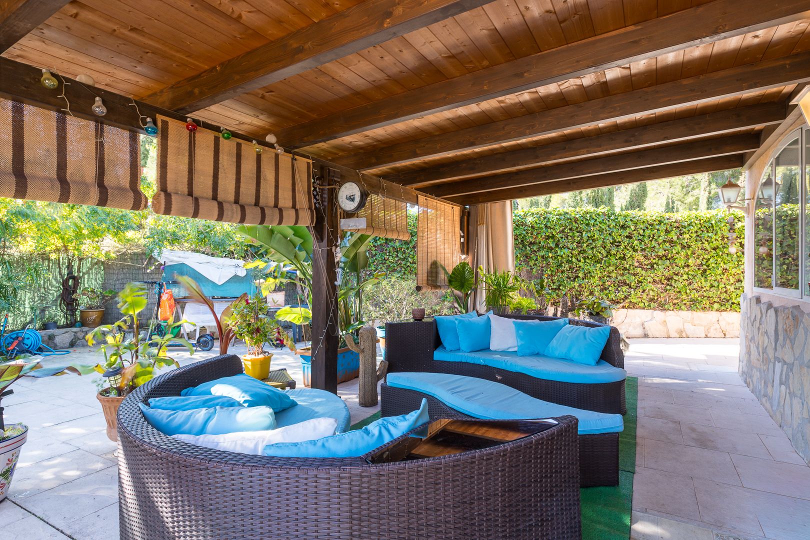 Covered terrace featuring a dark wood pergola, curved rattan lounge furniture with turquoise cushions, and natural bamboo blinds overlooking the garden.