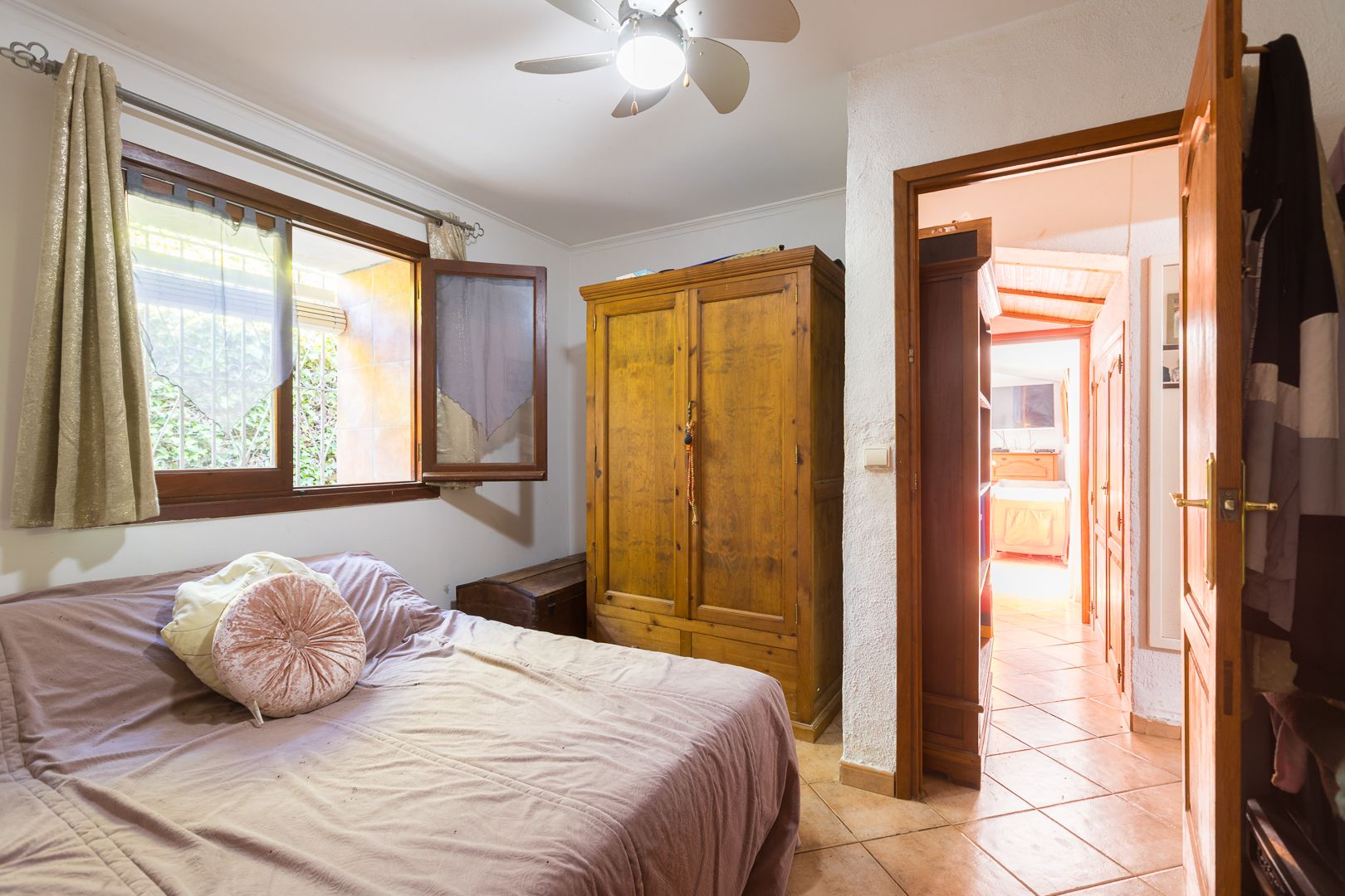 Cozy bedroom featuring a solid wood wardrobe, terracotta tile flooring, and a ceiling fan. A wooden-framed window provides natural light and a view of the exterior.