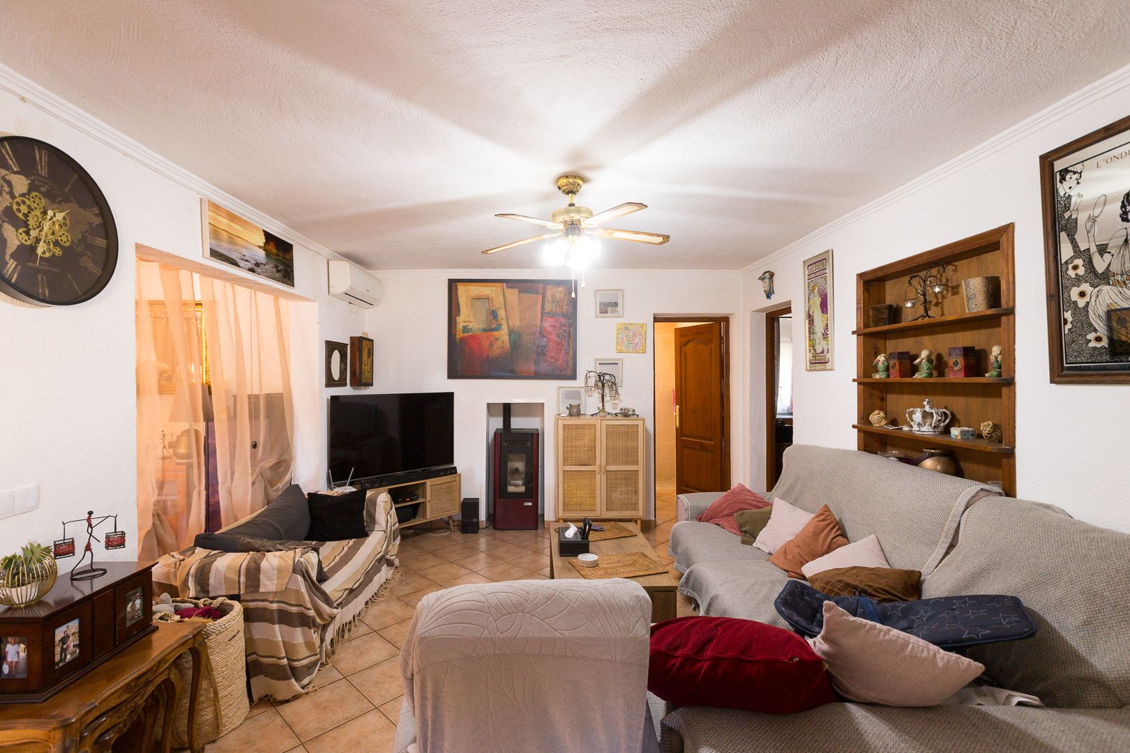Cozy living room featuring terracotta floor tiles, a pellet stove, air conditioning, and a ceiling fan. The space includes built-in wooden shelving and decorative wall art.