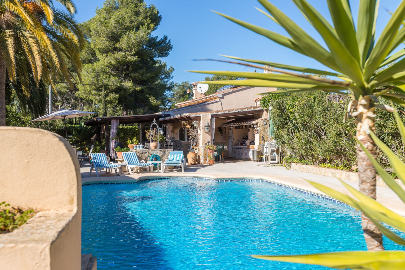 Heated swimming pool with solar panels on the roof, featuring a spacious sun terrace, loungers, and an adjacent covered summer kitchen.