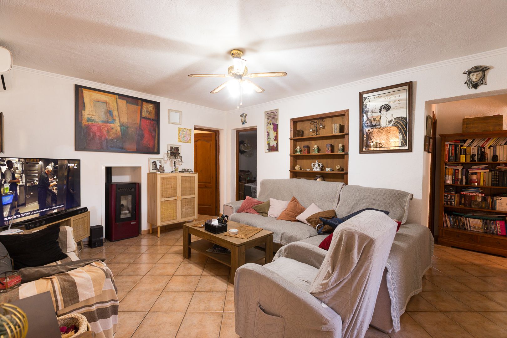 Living room featuring terracotta-style floor tiles, a pellet stove, ceiling fan, and built-in wooden shelving for a functional and warm atmosphere.