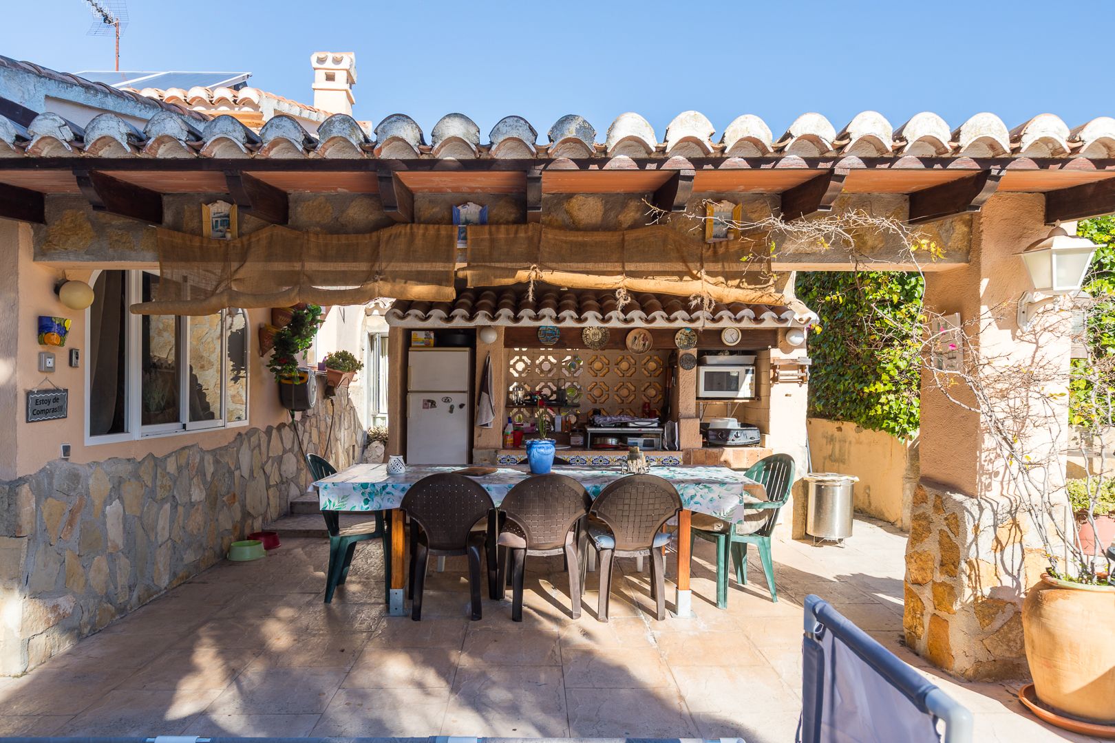 Outdoor summer kitchen and dining area under a wooden beam porch, featuring natural stone walls and a fully equipped preparation zone.
