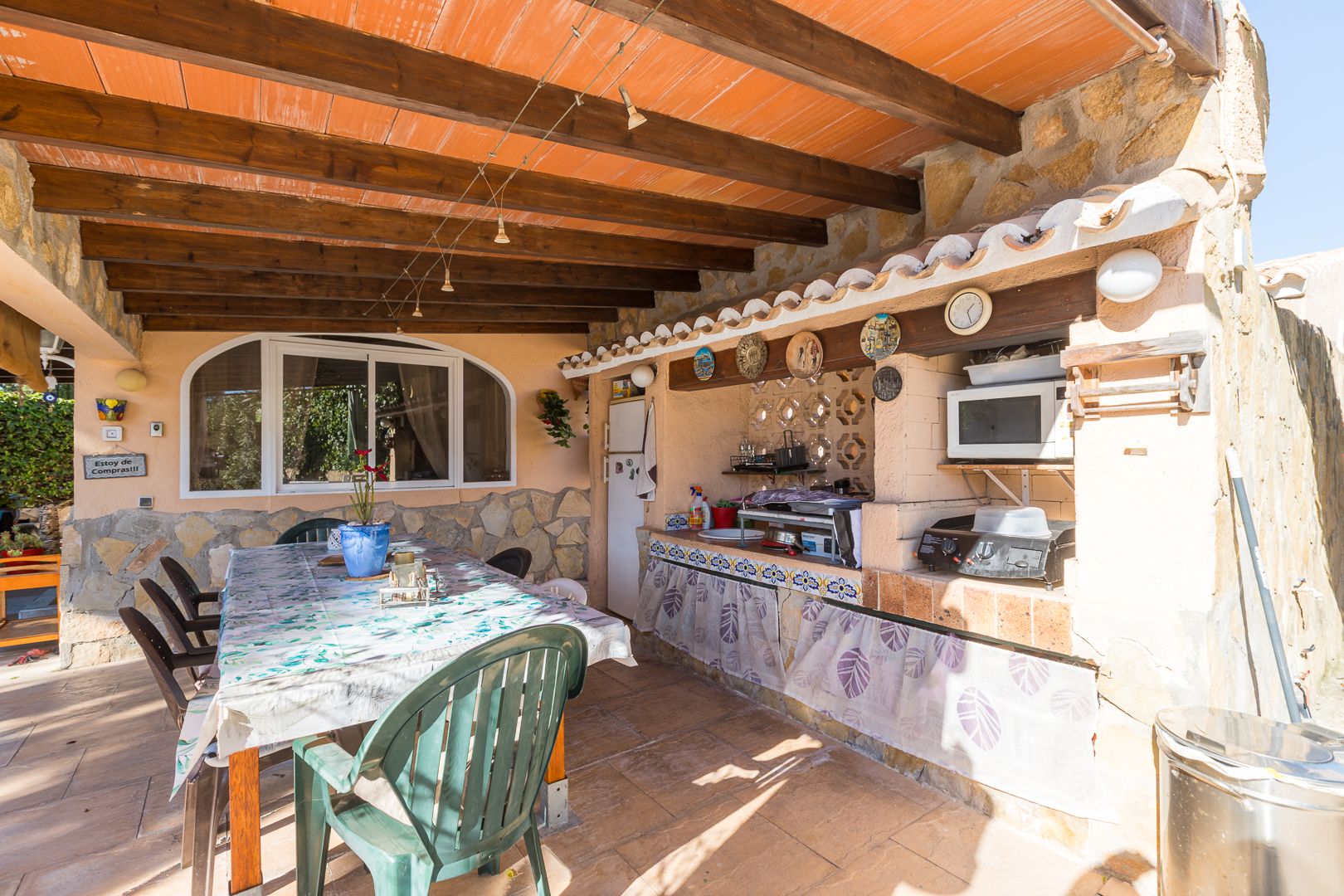 Outdoor summer kitchen and dining space under a rustic wooden beam ceiling, featuring stone walls and a built-in barbecue area.