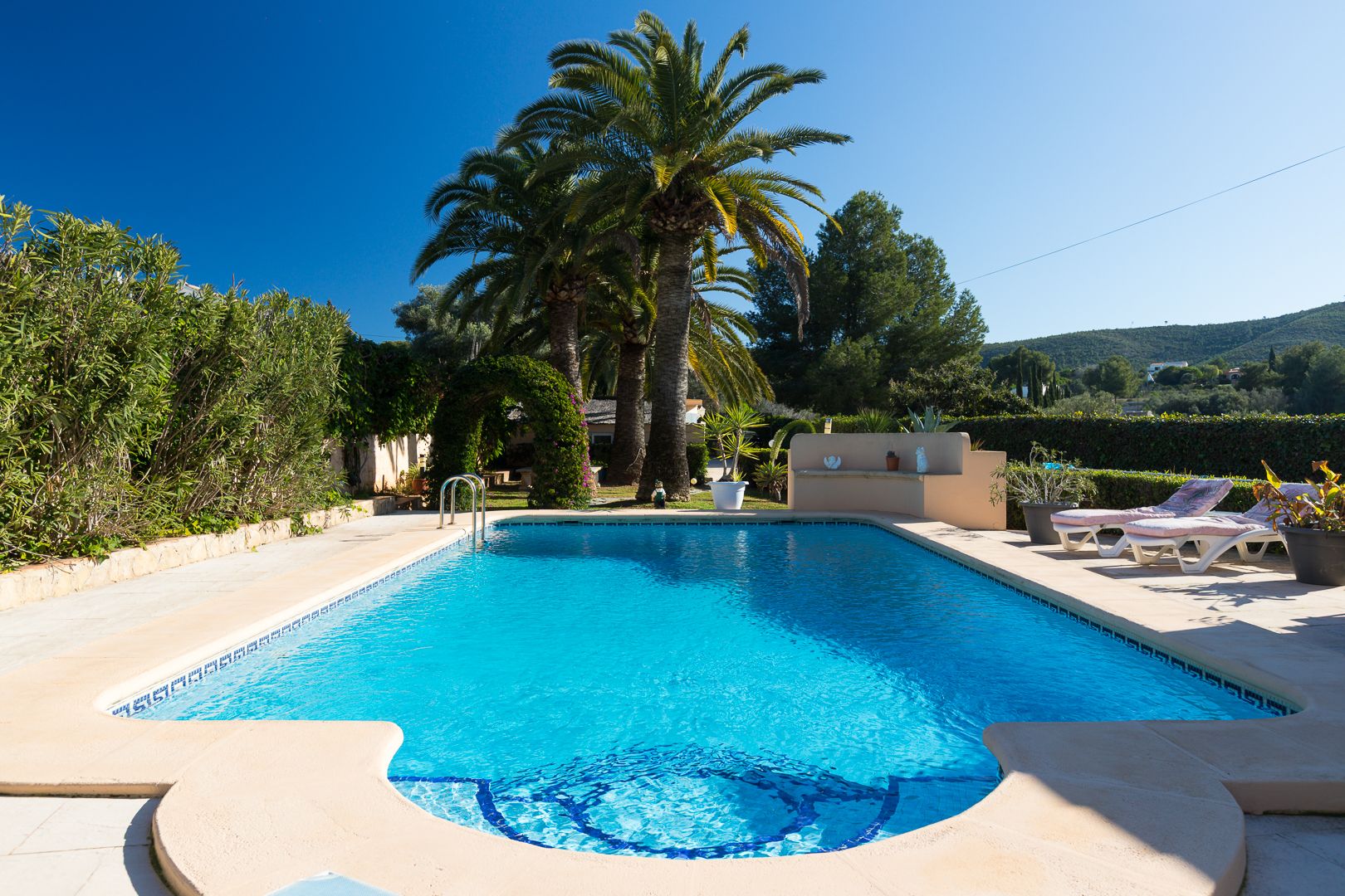 Rectangular swimming pool featuring integrated steps, surrounded by a stone sun terrace, mature palm trees, and views of the Mediterranean landscape.