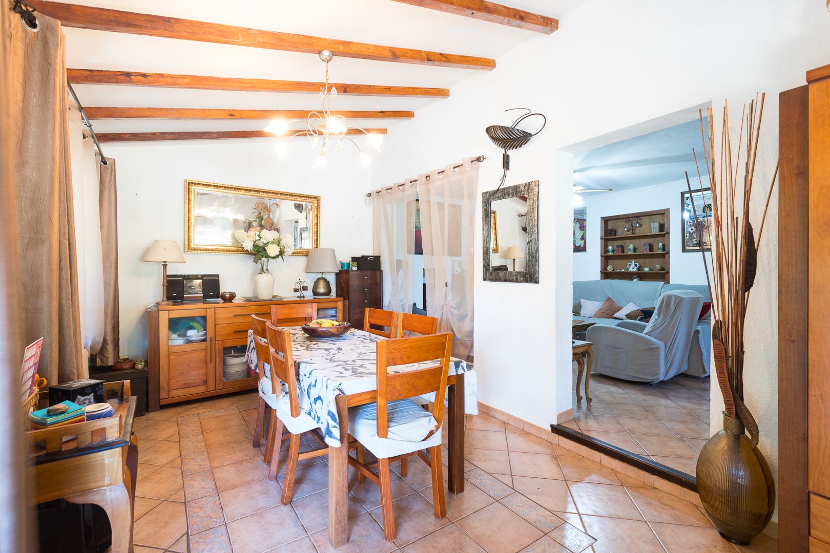 Rustic dining area featuring exposed wooden ceiling beams, terracotta floor tiles, and solid wood furniture with a view into the adjacent living room.