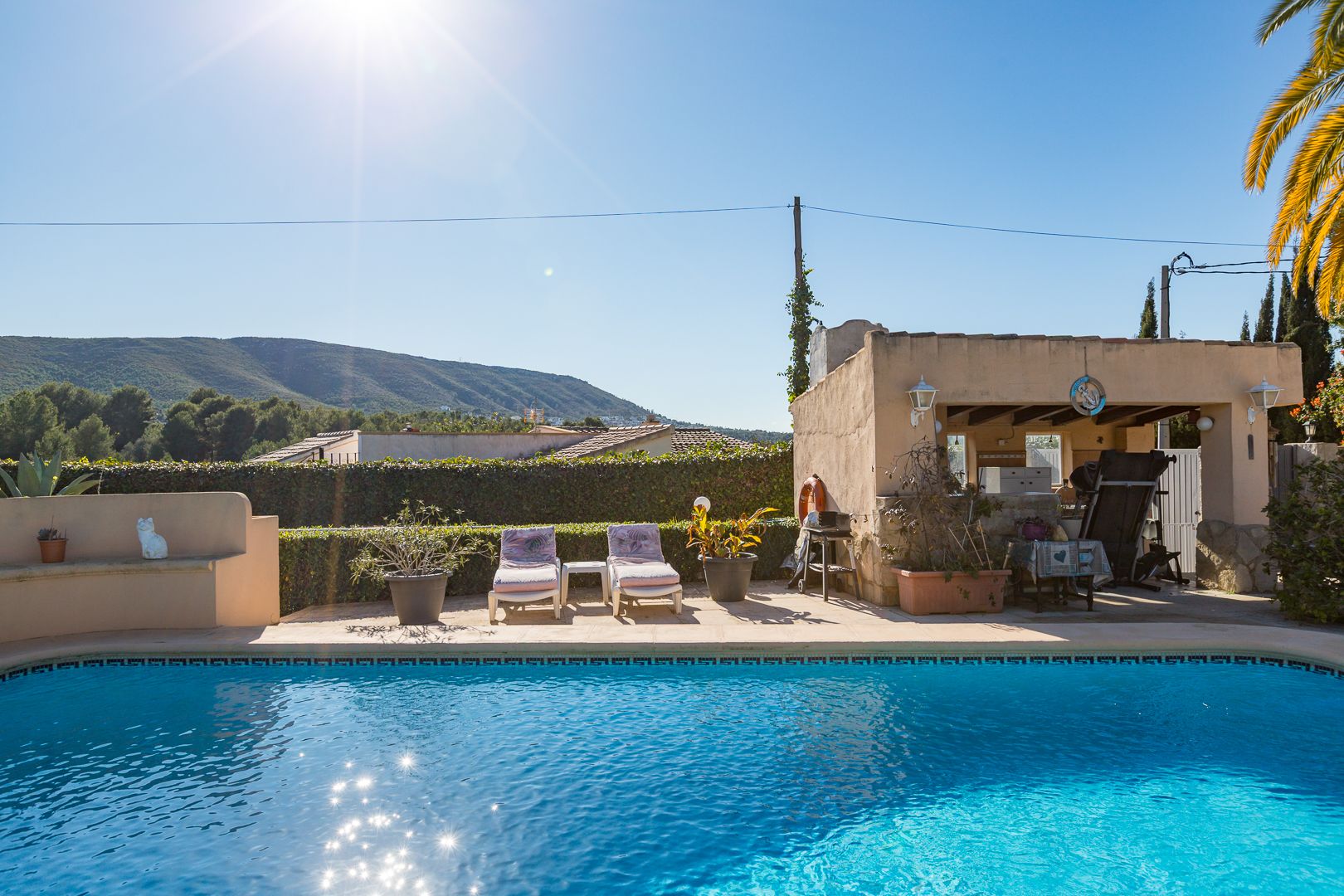 Tiled swimming pool featuring a sun terrace with loungers and a covered outdoor kitchen, set against a backdrop of mountain views and clear skies.