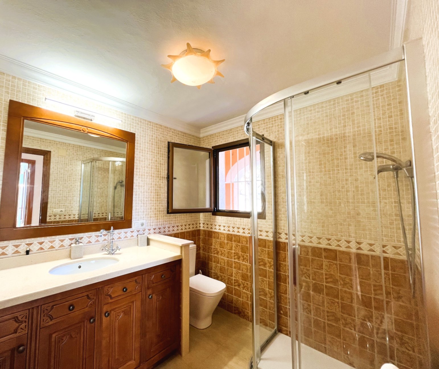 Bathroom featuring a carved wooden vanity, integrated sink, corner shower with curved glass doors, and earth-toned tiled walls with natural light.