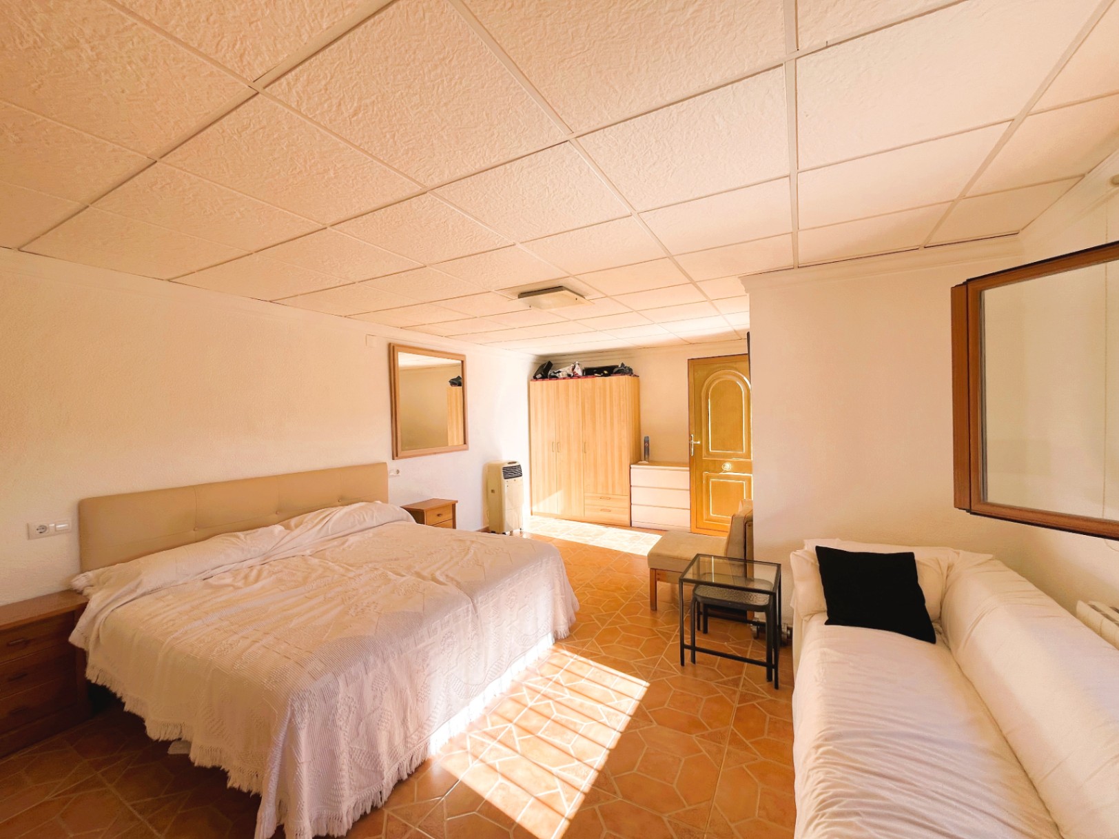 Bedroom featuring terracotta tile flooring, a modular drop ceiling, and a seating area. Natural light highlights the wooden furniture and spacious layout.